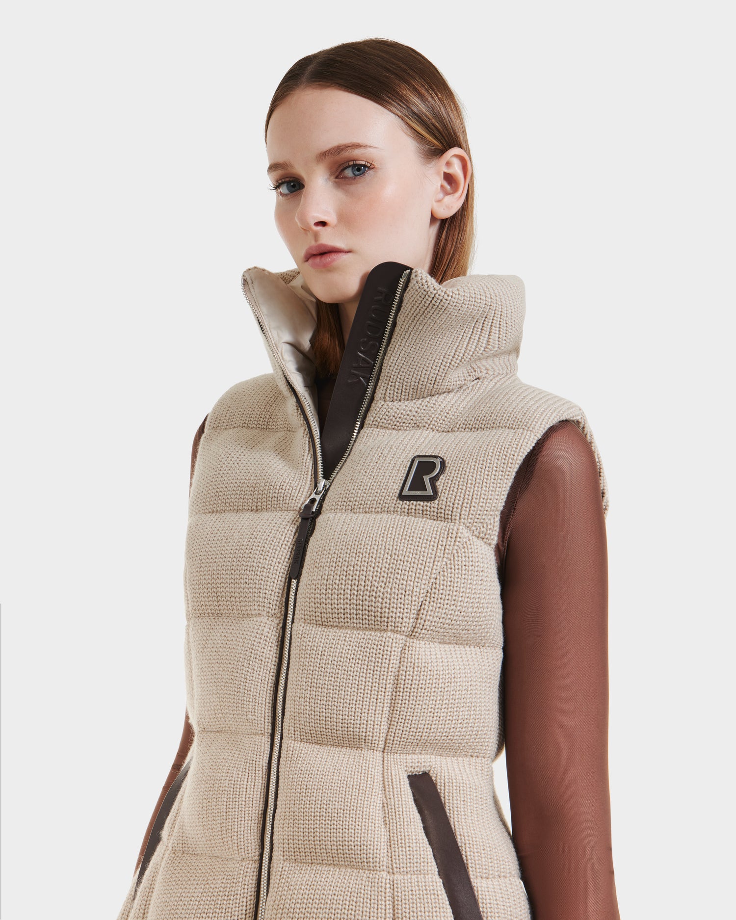 A woman wears the Rudsak CASSIDY - 8125622 Vanilla beige quilted fitted down vest with a high collar, front zipper, brown trim, and black R logo on the chest, styled over a sheer brown long-sleeve top.