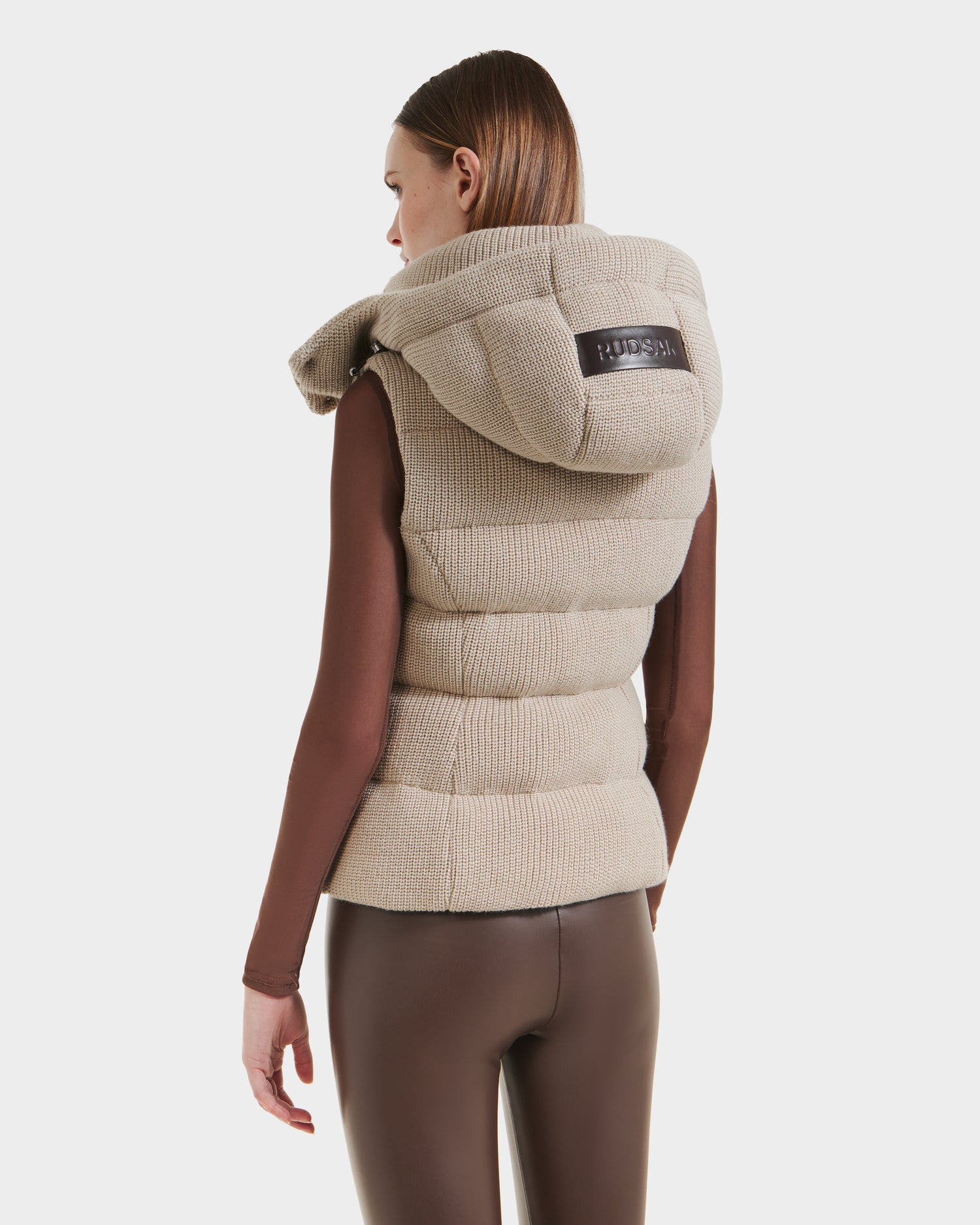 A woman is shown from the back in a Rudsak CASSIDY - 8125622 Vanilla womens fitted down vest featuring lightweight warmth and a black label on the removable hood, paired with a brown long-sleeve top and fitted pants against a white background.