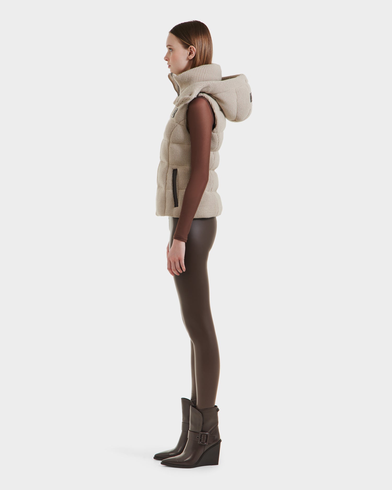 A woman stands in profile wearing the Rudsak CASSIDY - 8125622 Vanilla women’s fitted down vest with a large removable hood, styled with brown leggings, a long-sleeve brown top, and dark brown heeled boots against a plain white background.