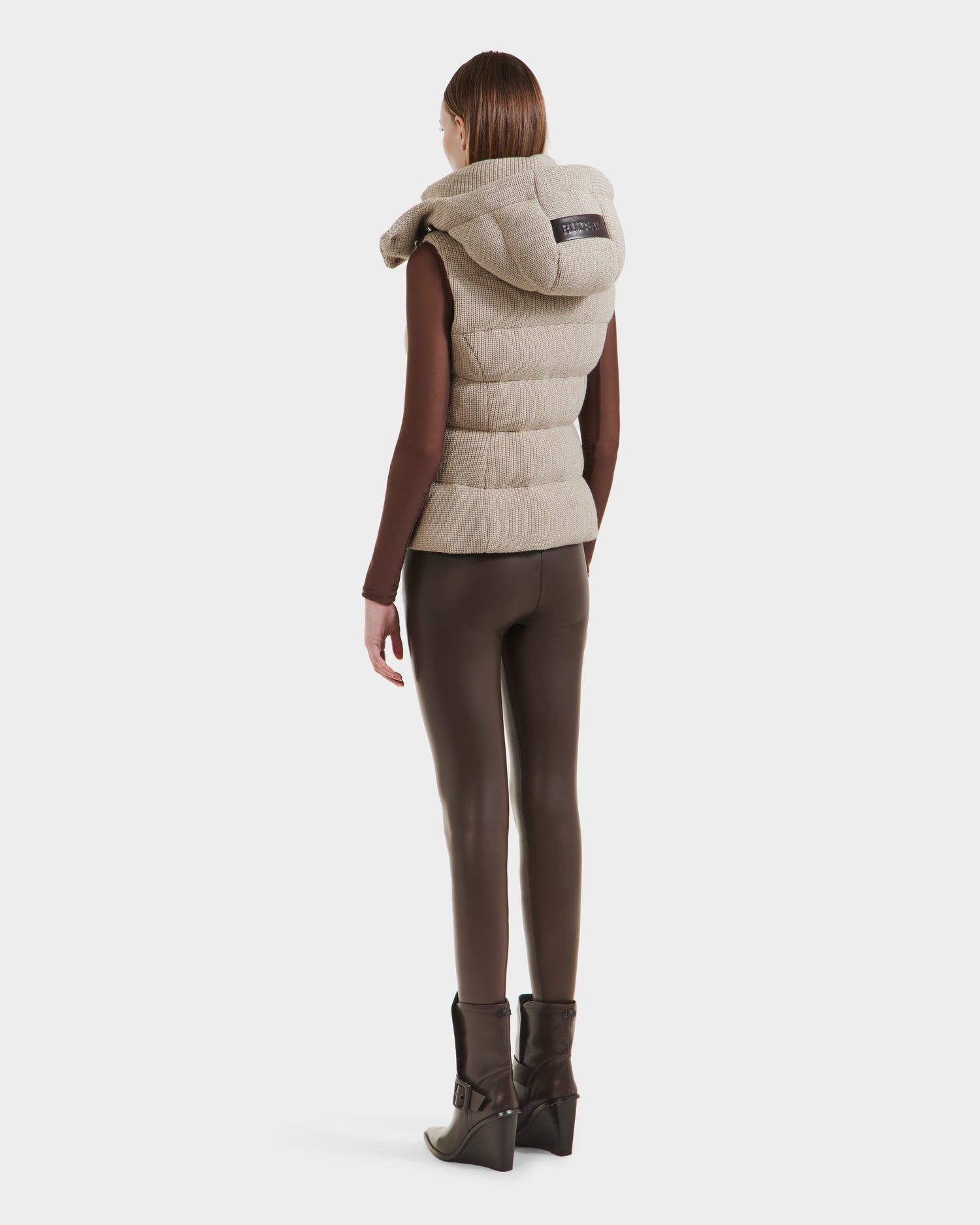A woman faces away against a white background, wearing the Rudsak CASSIDY - 8125622 Vanilla women’s fitted down vest with a large collar, styled with a brown long-sleeve top, fitted pants, and heeled boots.