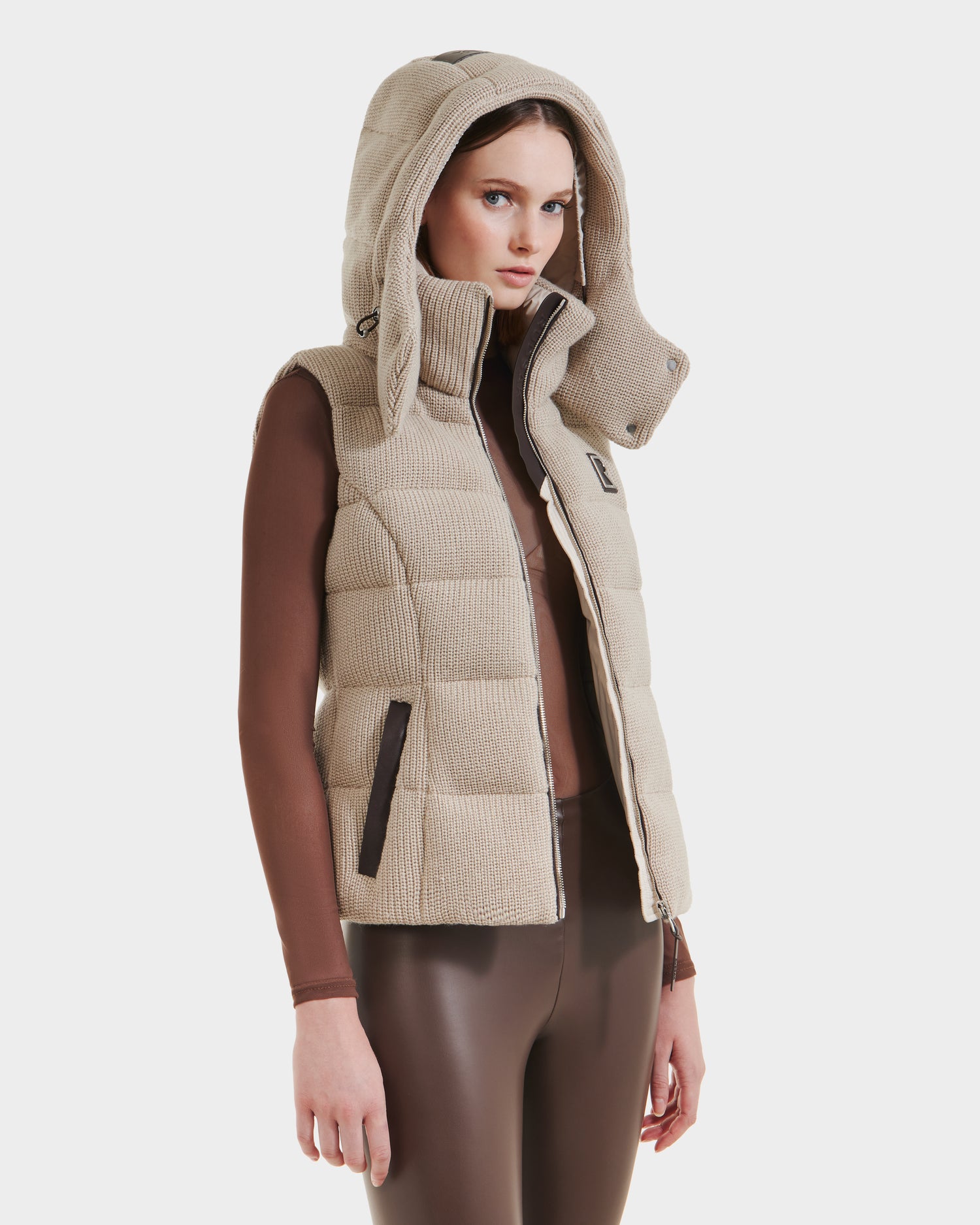 A woman wears the Rudsak CASSIDY - 8125622 Vanilla women’s fitted down vest with a removable hood, layered over a brown long-sleeve top and brown leggings, offering lightweight warmth against a plain white background.