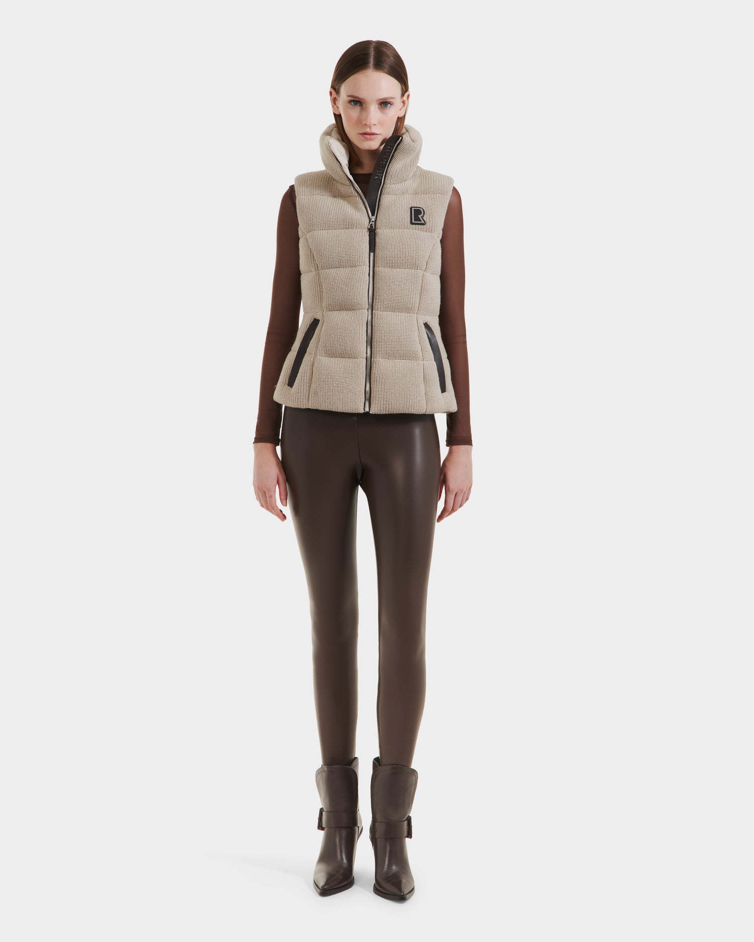 A woman faces forward against a plain white background, wearing the Rudsak CASSIDY - 8125622 Vanilla women’s fitted down vest over a brown long-sleeve top, paired with brown shiny leggings and heeled ankle boots.