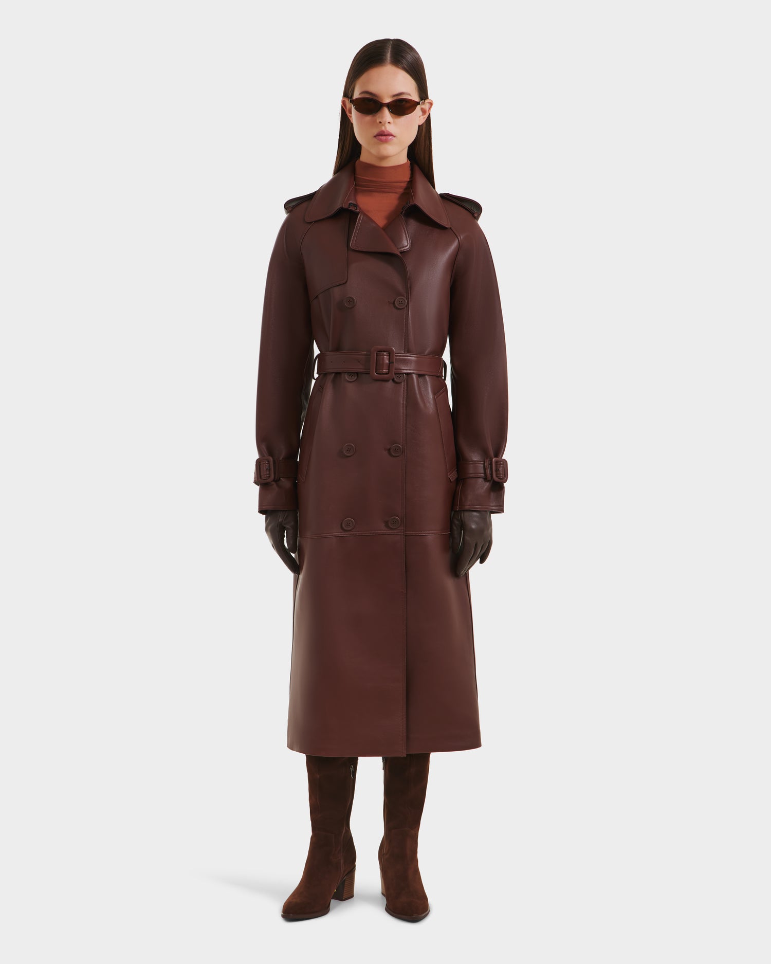 A woman in a Rudsak CASSIE L-B - 8125553 Rust long trench coat, belted at the waist, with gloves, boots, sunglasses, and a turtleneck stands facing forward against a plain white background.