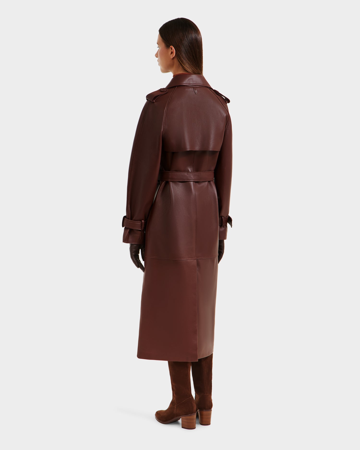 A woman with loose, straight dark hair faces away, wearing the Rudsak CASSIE L-B - 8125553 Rust bonded leather trench coat with belt. She pairs it with brown gloves and knee-high boots, standing against a plain light background.