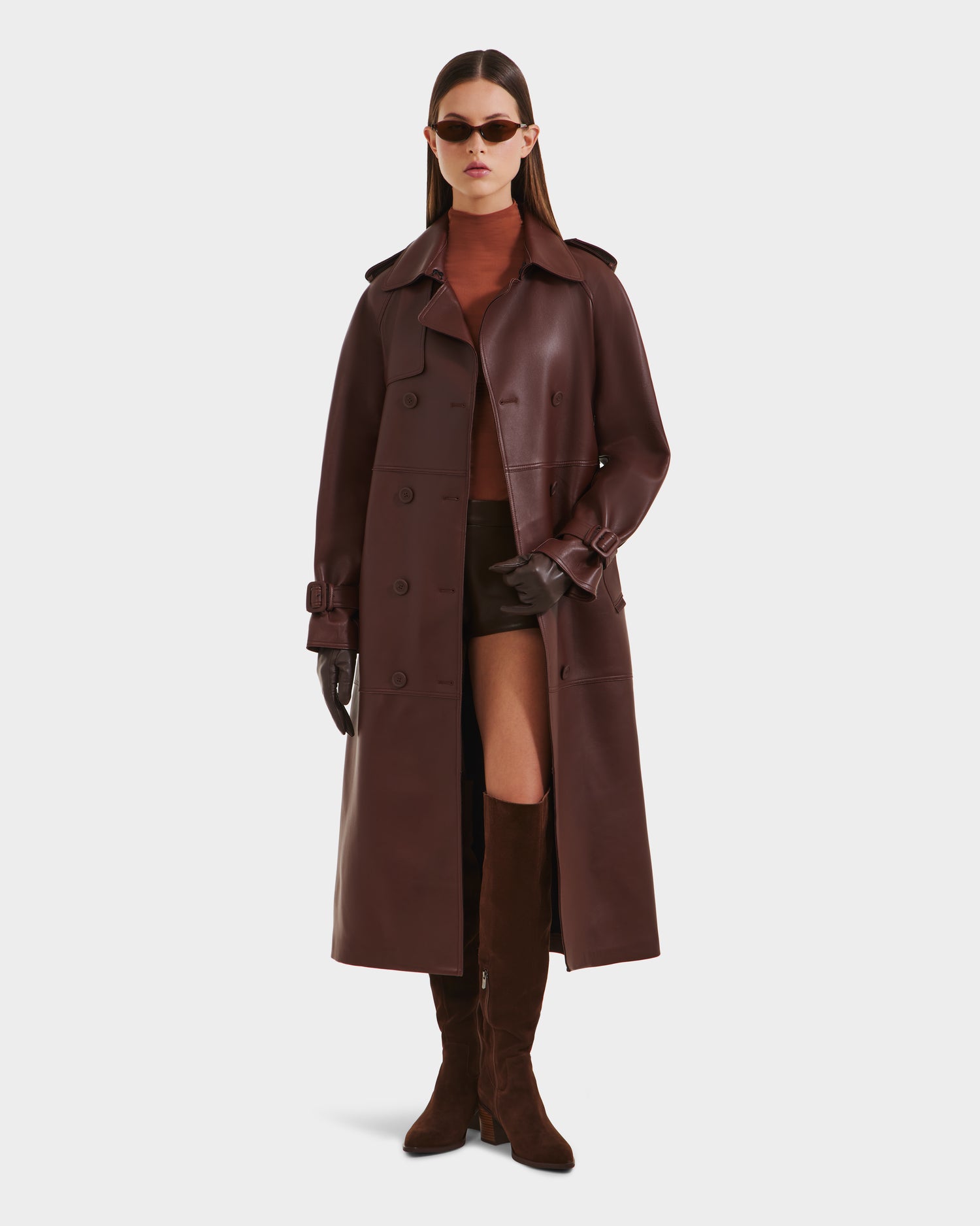 A woman models the Rudsak CASSIE L-B - 8125553 Rust coat with brown boots, gloves, sunglasses, and a rust-colored top, standing hands-in-pockets against a plain white background.