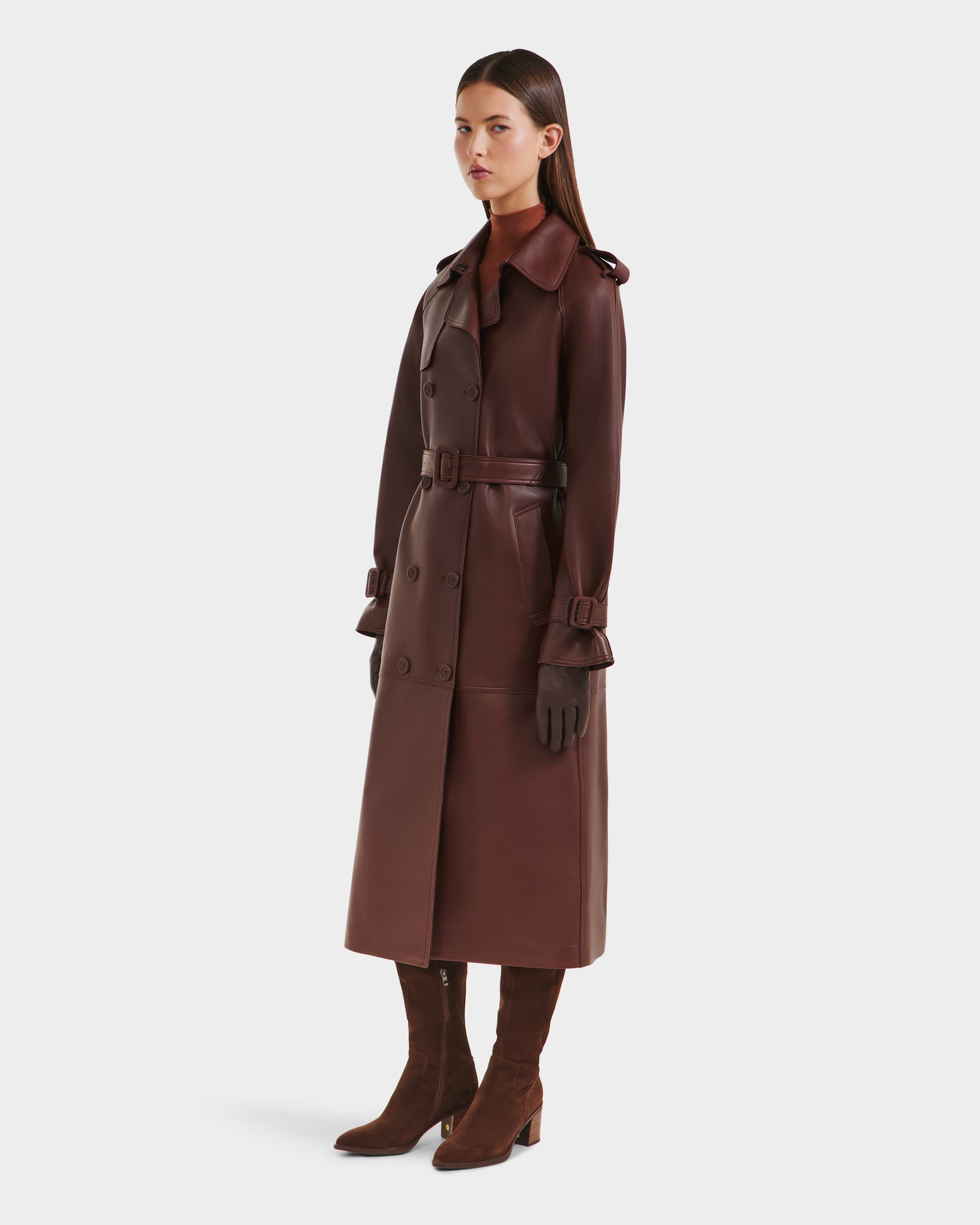 A woman with straight dark hair and a neutral expression stands facing forward against a plain background, wearing the Rudsak CASSIE L-B - 8125553 Rust bonded leather trench coat, dark gloves, and brown suede knee-high boots.