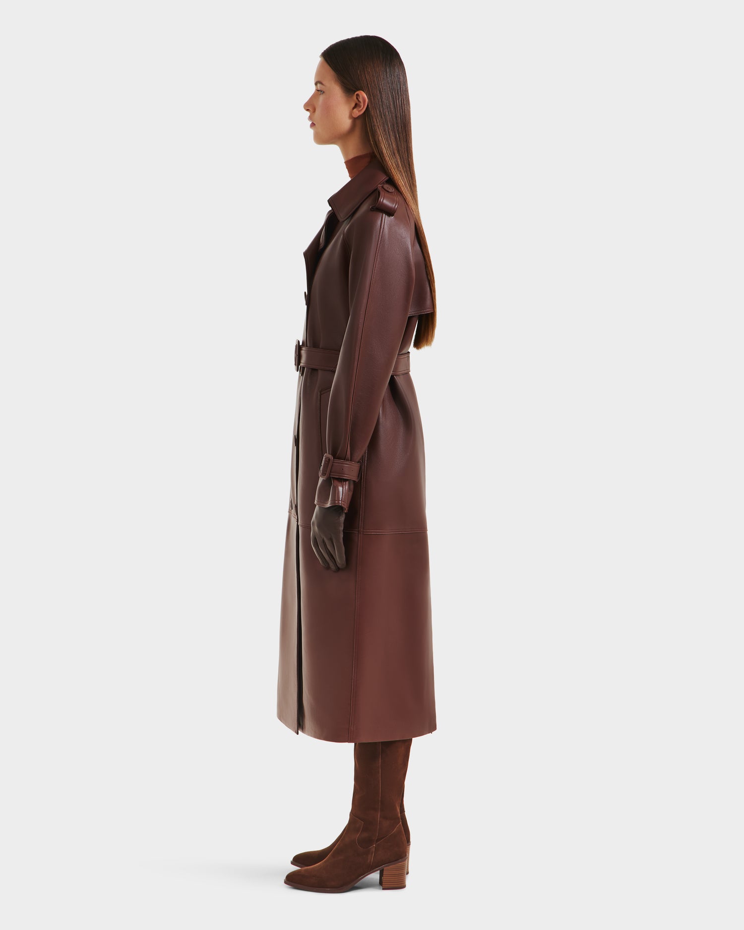 A woman with straight, long hair stands in profile against a plain white background, wearing the Rudsak CASSIE L-B - 8125553 Rust trench coat in bonded leather, styled with a belt, brown knee-high boots, and gloves.