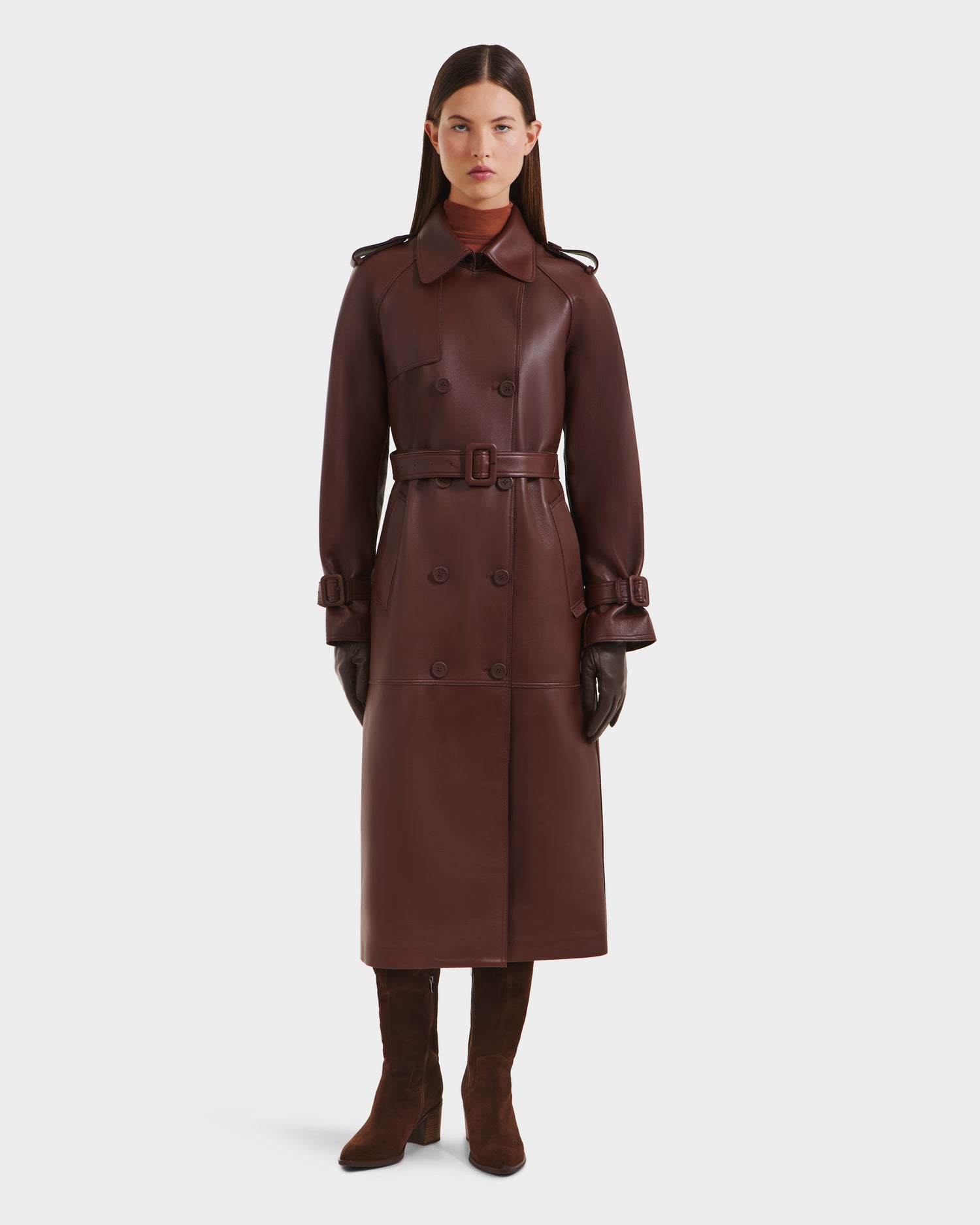 A woman faces forward wearing the Rudsak CASSIE L-B - 8125553 Rust long double-breasted trench coat in dark brown leather, with matching gloves and heeled boots, against a plain white background.