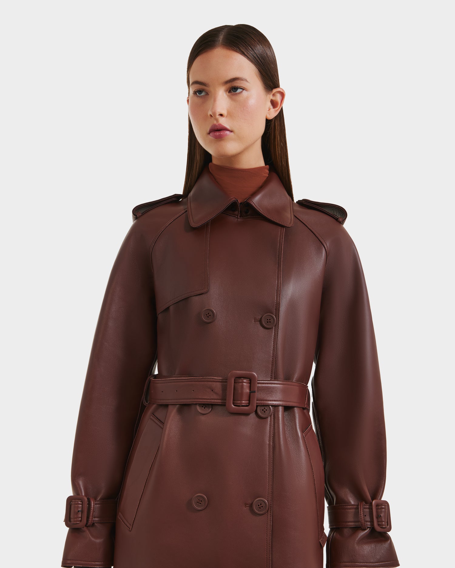 A woman with straight brown hair models the Rudsak CASSIE L-B - 8125553 Rust long double-breasted trench coat in dark brown bonded leather, featuring matching buttons and waist buckle, set against a plain white background.