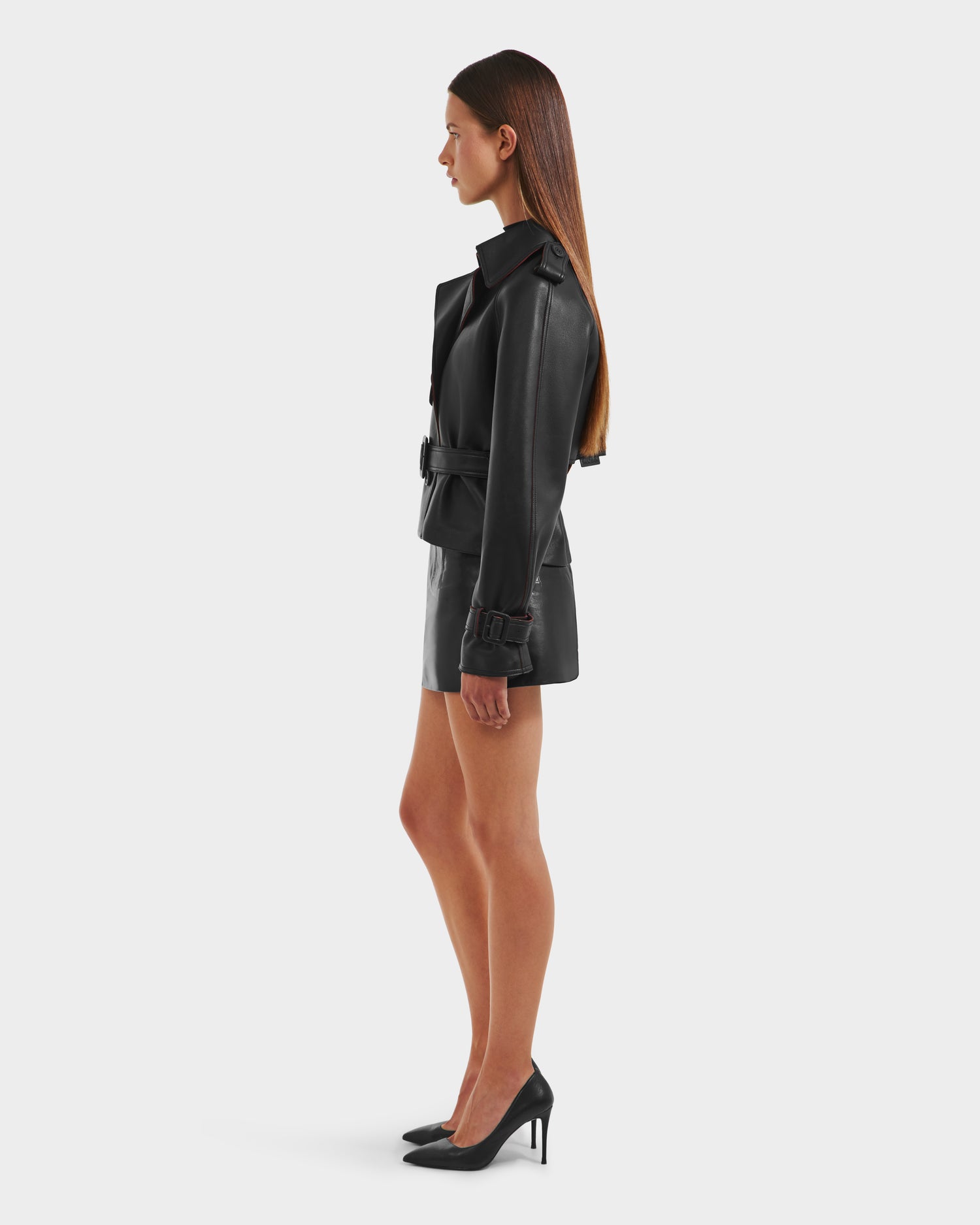 A woman with straight brown hair stands in profile against a plain white background, wearing the Rudsak CASSIE S-B - 8125554 Black belted bonded leather jacket, a matching mini skirt, and black high-heeled shoes.