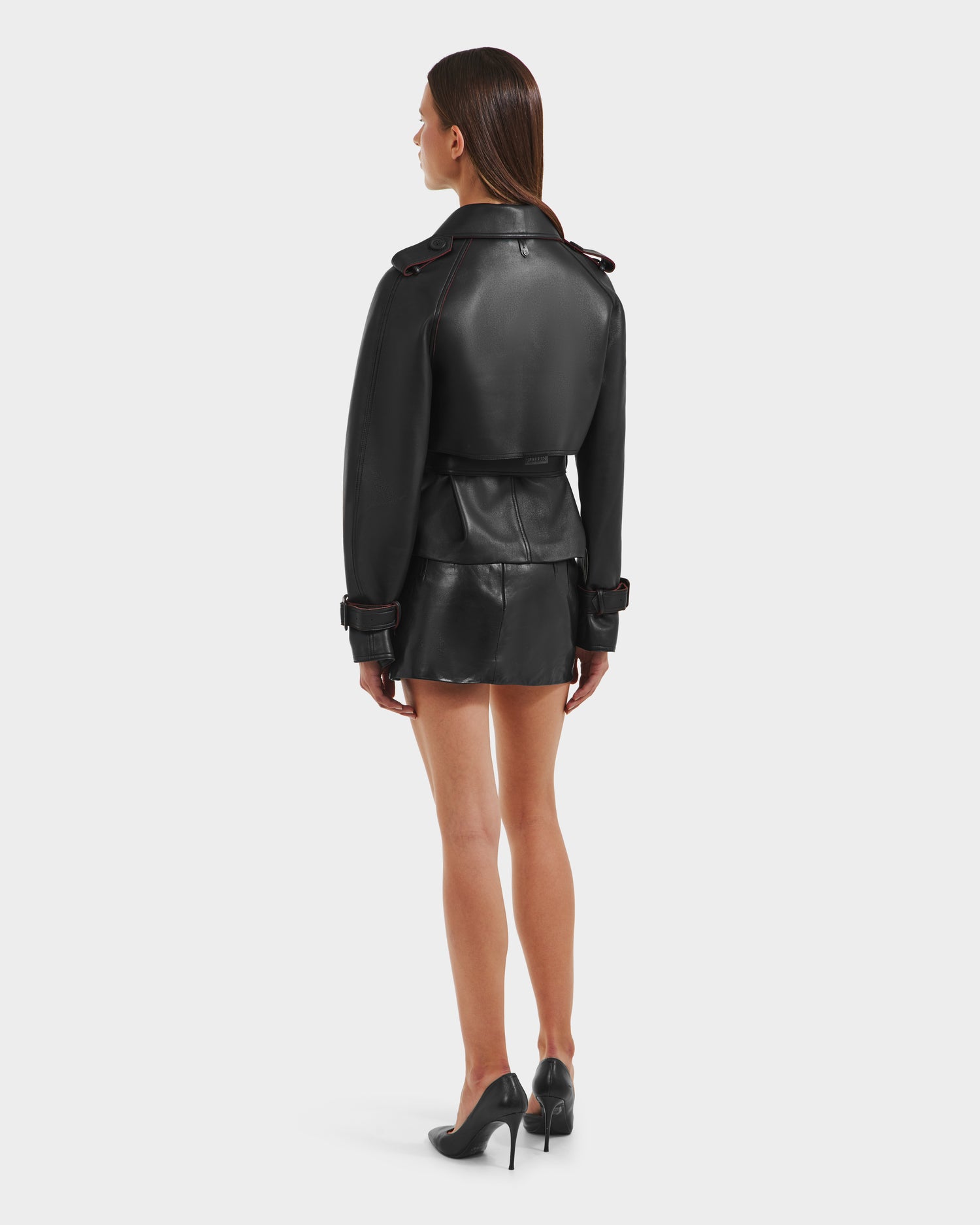 A woman with straight brown hair wears the Rudsak CASSIE S-B - 8125554 Black jacket and matching mini skirt with high heels, standing and facing away from the camera against a plain white background.