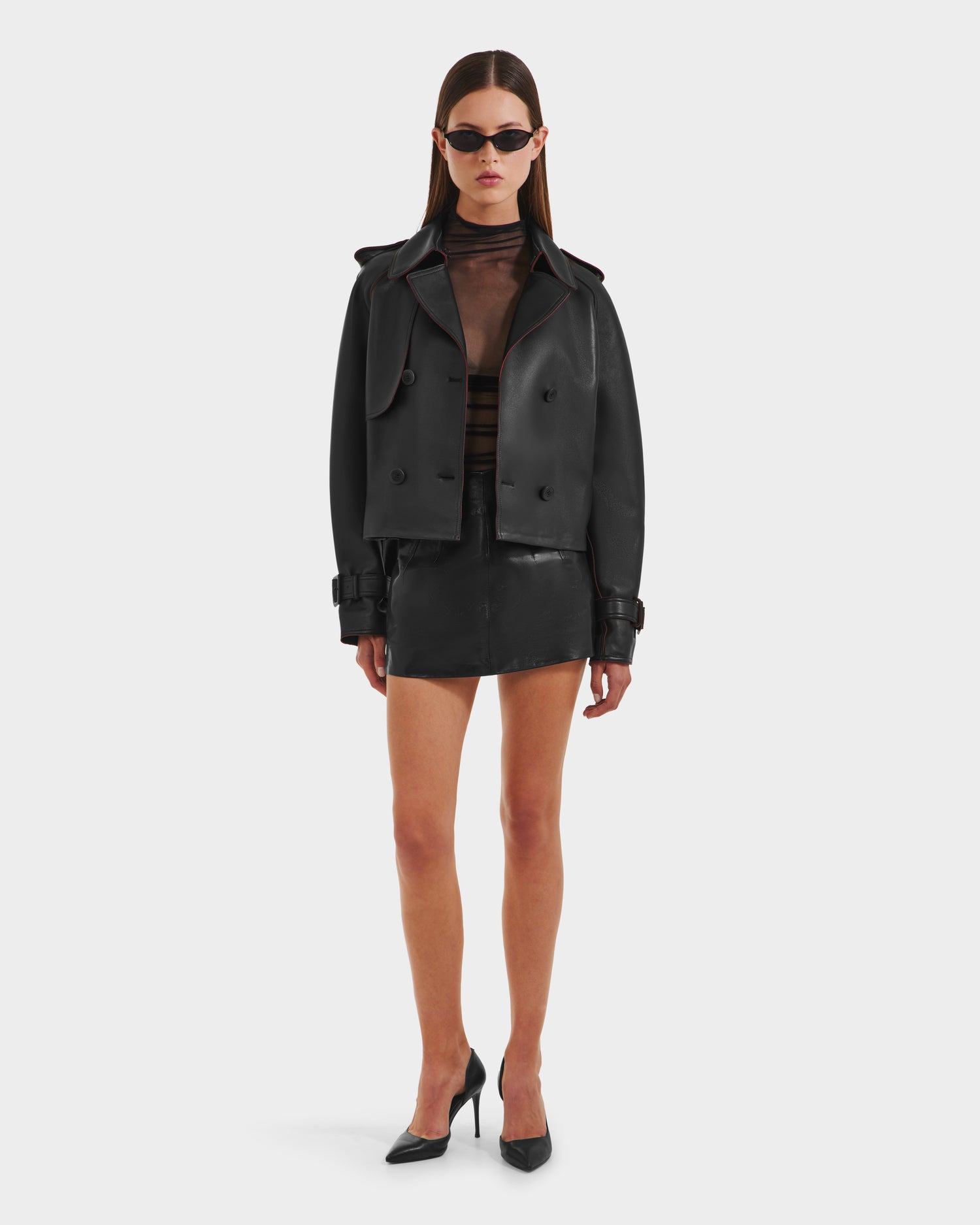 A woman wearing black sunglasses, a sheer black top, the Rudsak CASSIE S-B - 8125554 Black cropped trench coat, a bonded leather mini skirt, and black high-heeled shoes stands against a plain white background.