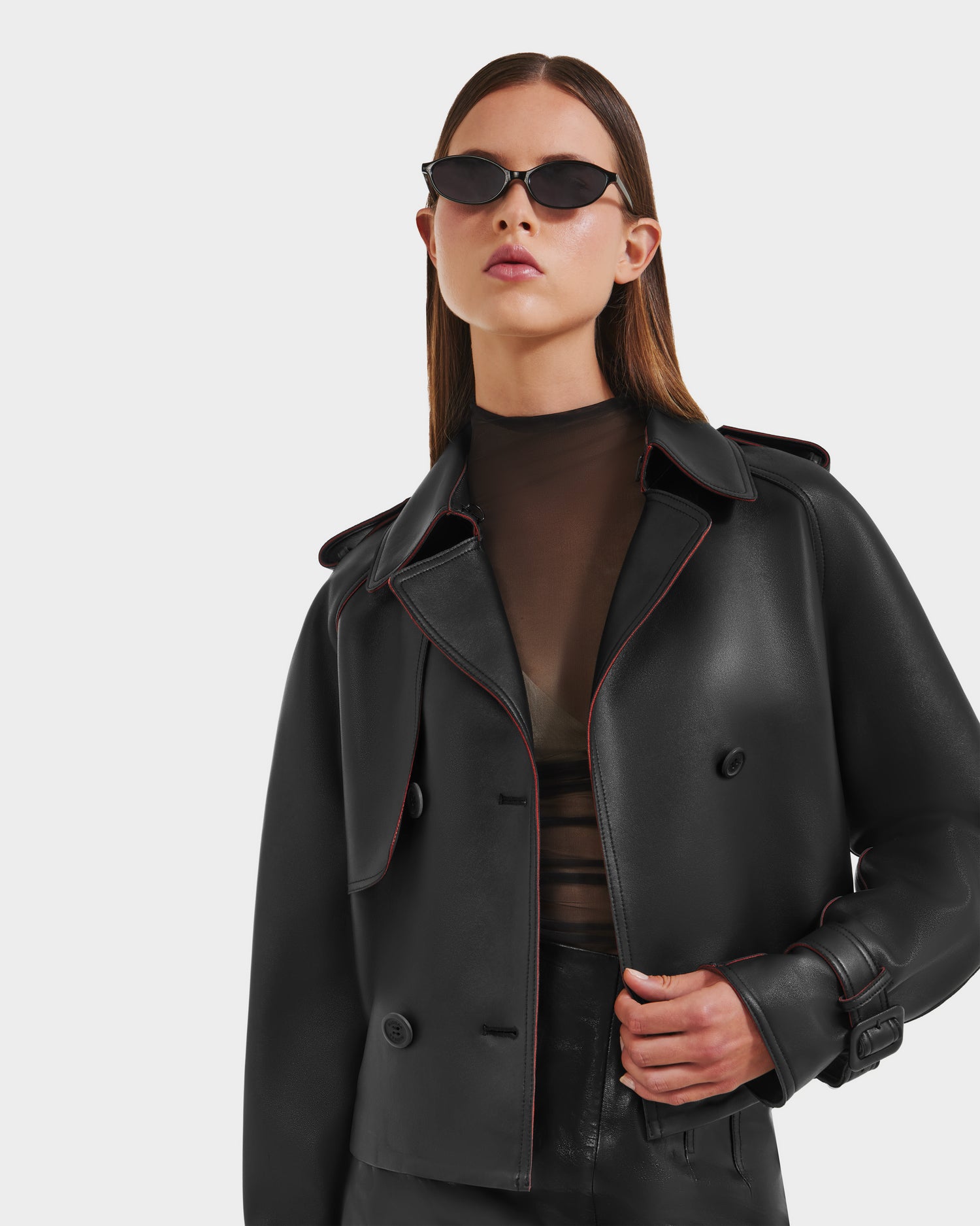 A woman in black cat-eye sunglasses, a sheer black top, and the Rudsak CASSIE S-B - 8125554 Black jacket with red piping stands against a white background, looking confidently at the camera.
