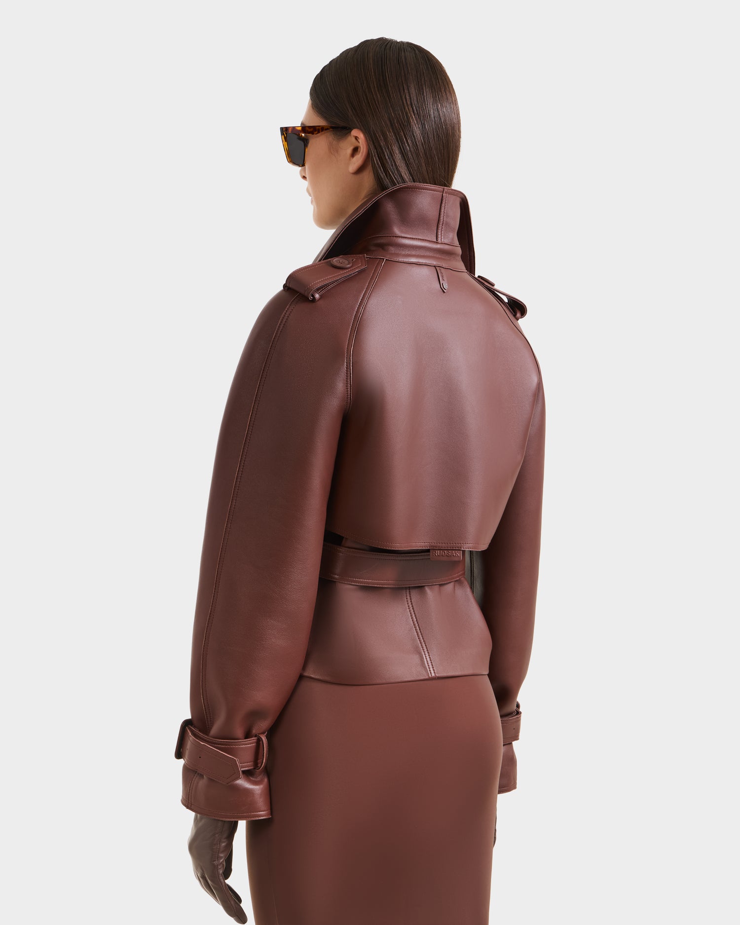 A person in sunglasses models the Rudsak CASSIE S-B - 8125554 Rust double-breasted bonded leather jacket with a belted waist and high collar, viewed from behind against a plain white background.
