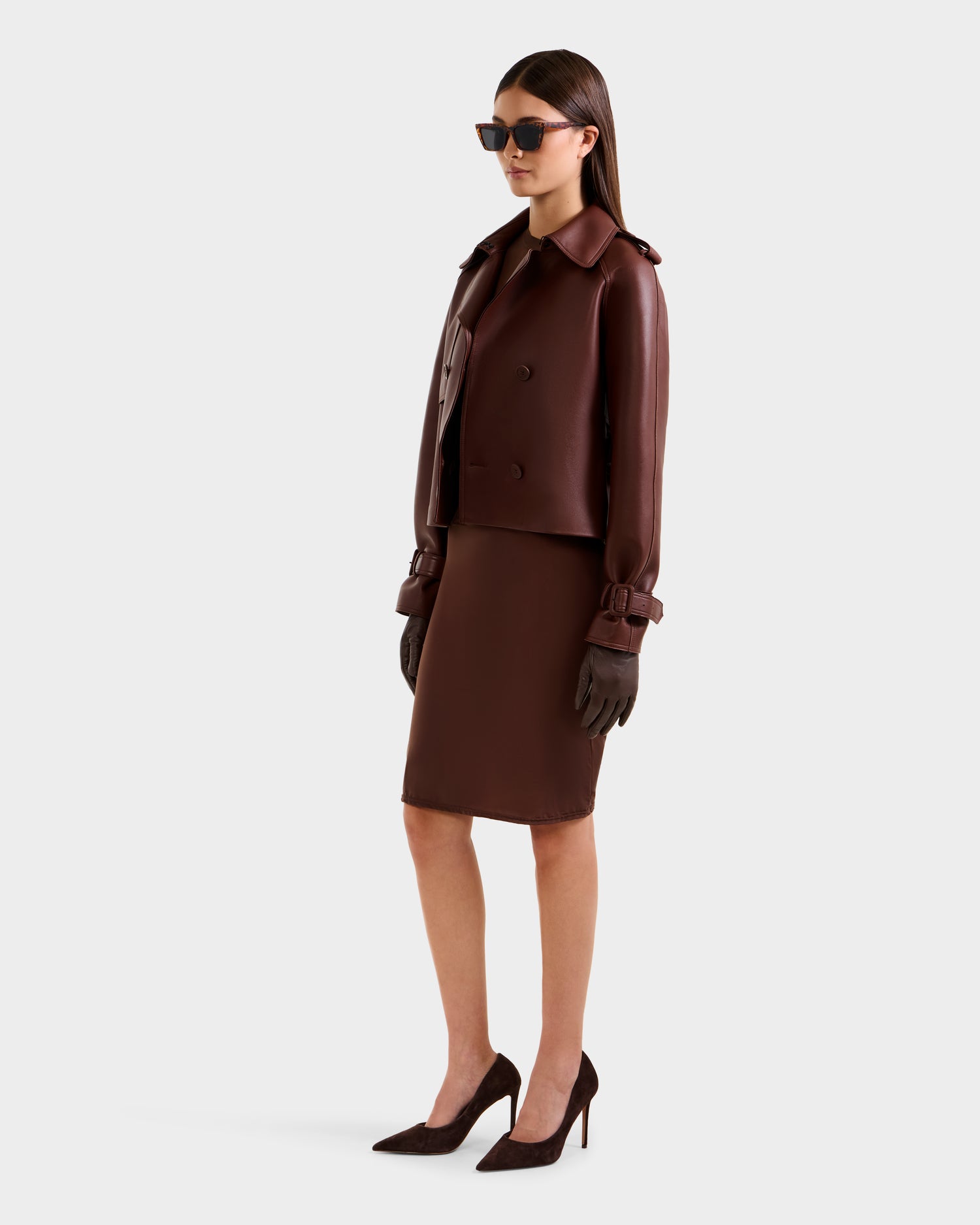 A woman in Rudsak's CASSIE S-B - 8125554 Rust trench coat with matching gloves, a brown knee-length skirt, sunglasses, and high heels stands against a plain light background.