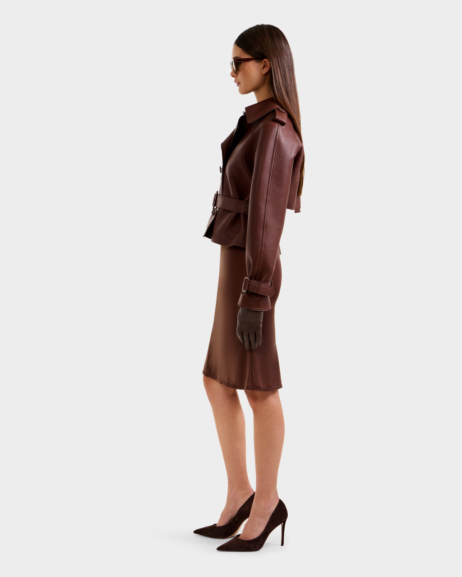 A woman in profile wears the Rudsak CASSIE S-B - 8125554 Rust double-breasted, belted bonded leather jacket with a matching brown skirt, pointed heels, gloves, and sunglasses against a plain light gray background.