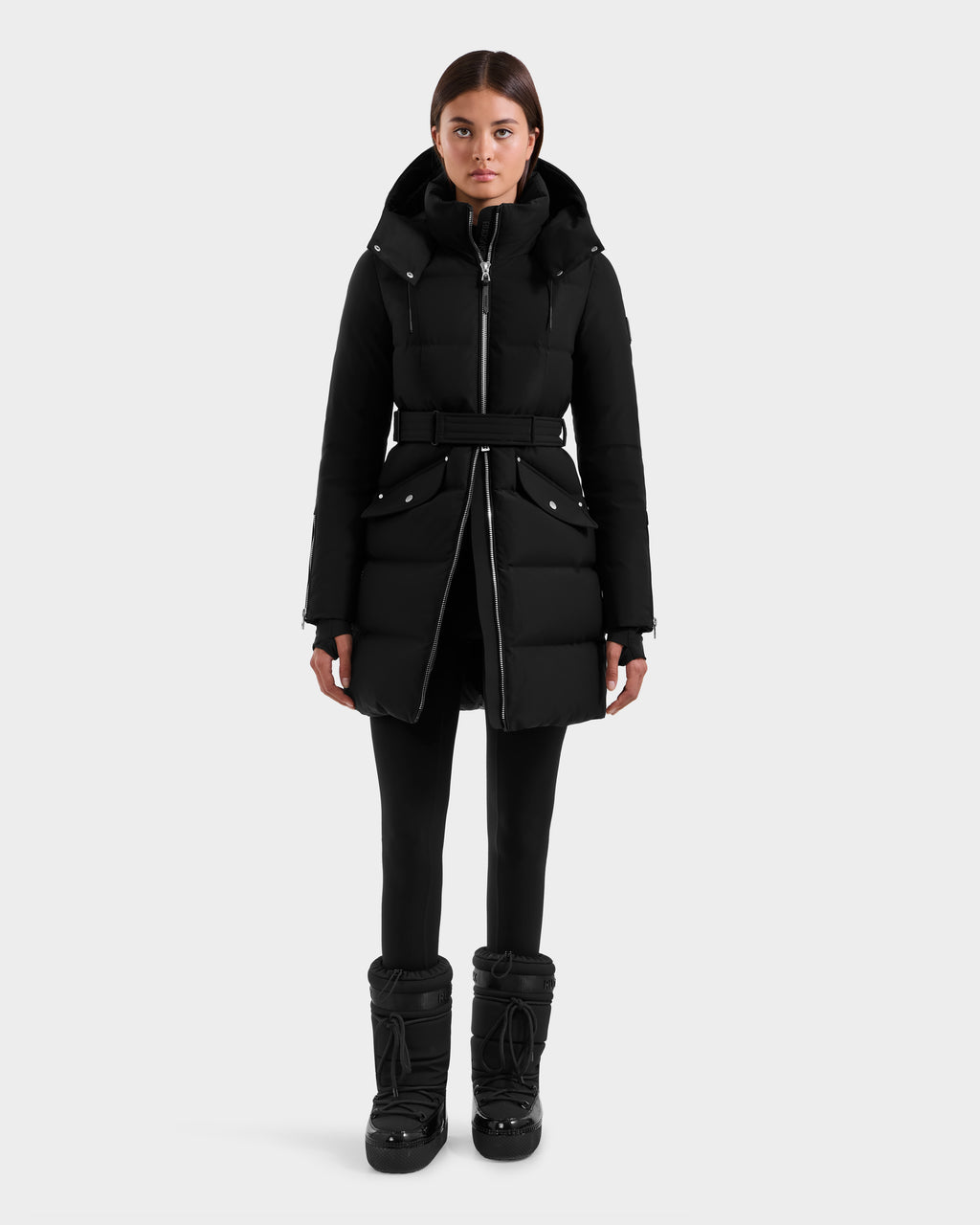 A person faces forward, wearing the Rudsak CAT H - 8125588 Black women's fitted down puffer with large front pockets, paired with black leggings and bulky black winter boots, against a plain light gray background.