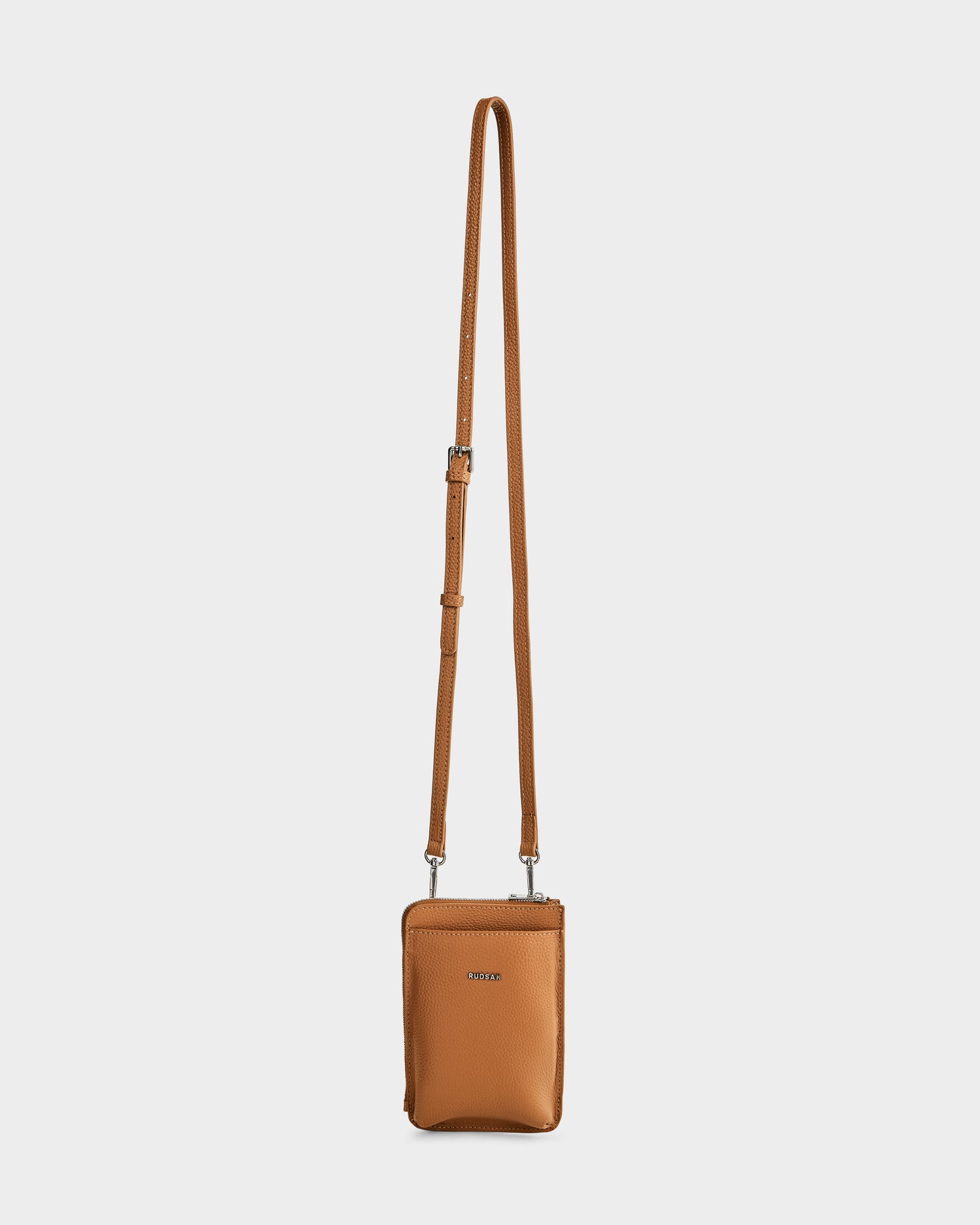 A small, rectangular taupe women's leather crossbody bag by Rudsak (CELYN - L325514) with an adjustable strap, silver hardware, pebbled leather, zip closure, and the Rudsak logo on the front, shown against a plain white background.