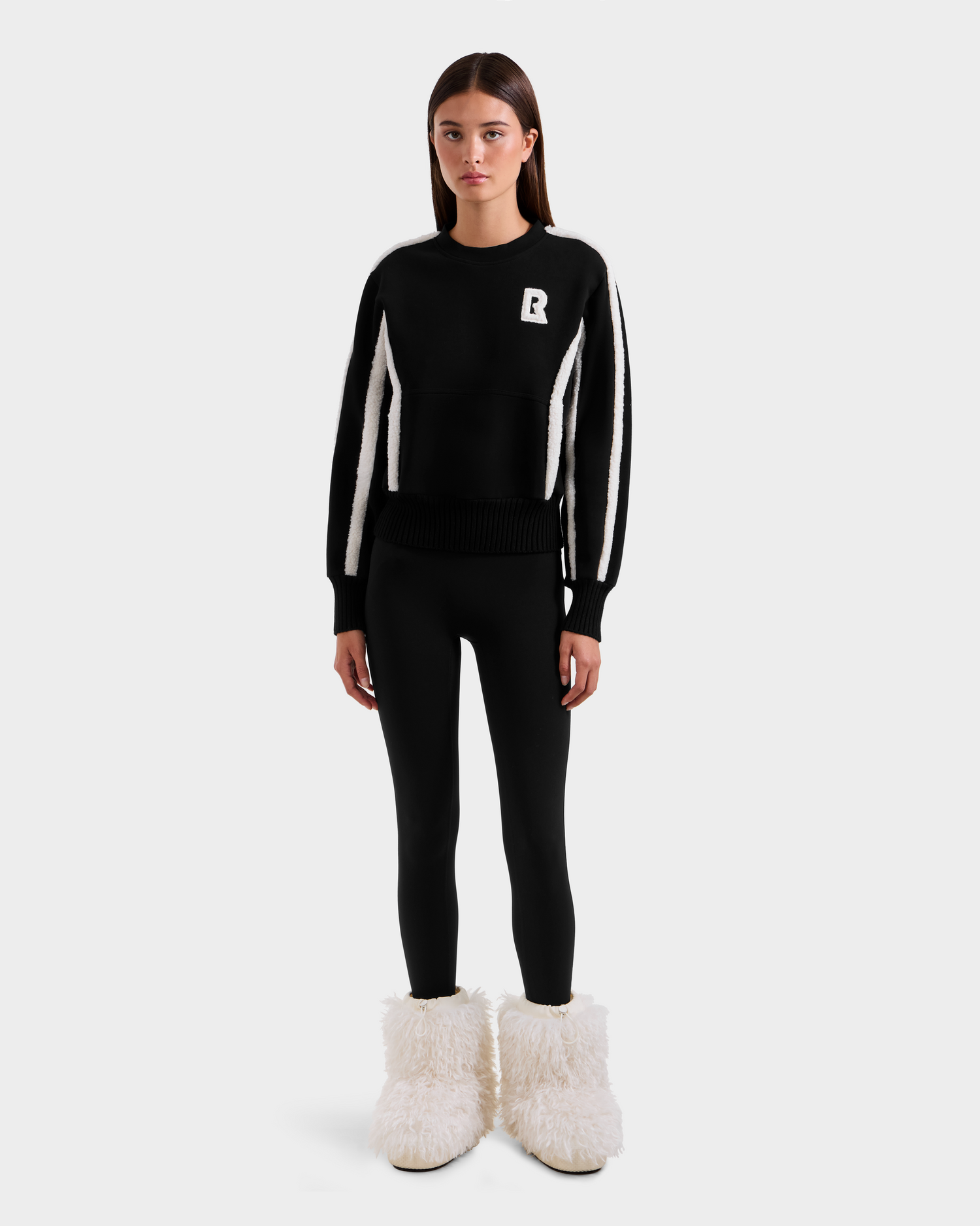 A woman faces forward in a Rudsak CHANA - 8525656 Black women’s crew neck sweatshirt with white sleeve stripes and a white R on the chest, paired with black leggings and fluffy faux shearling-trimmed boots, against a plain white background.