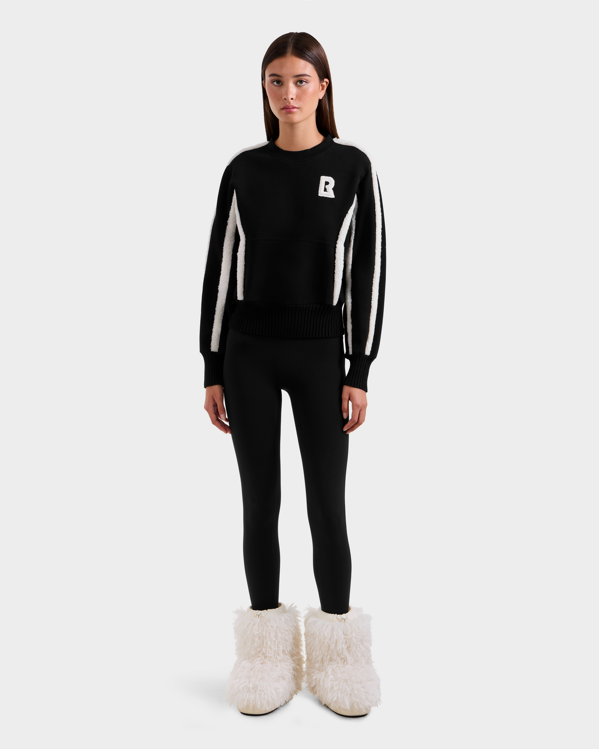 A woman faces forward in a Rudsak CHANA - 8525656 Black women’s crew neck sweatshirt with white sleeve stripes and a white R on the chest, paired with black leggings and fluffy faux shearling-trimmed boots, against a plain white background.
