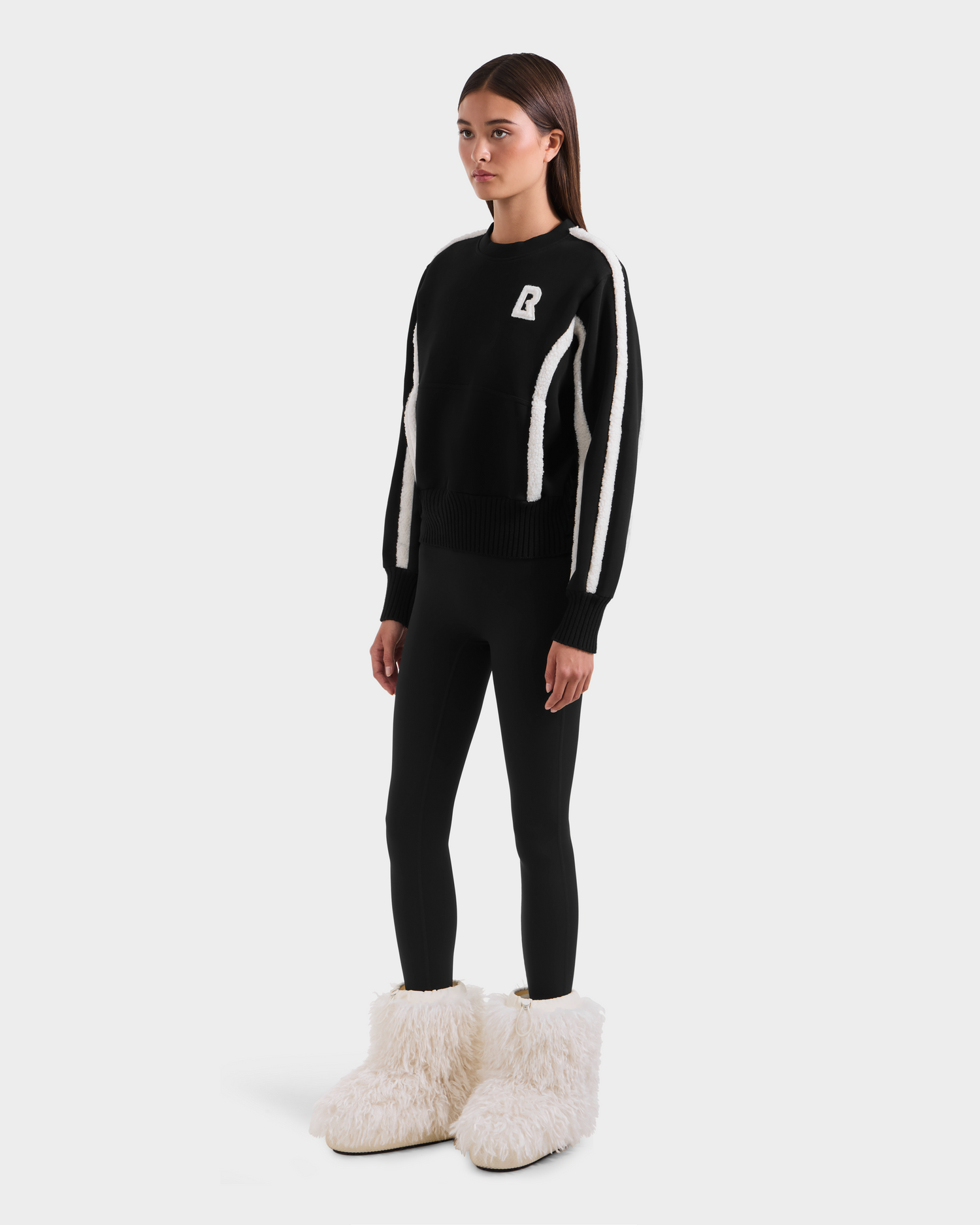 A woman stands against a plain background wearing Rudsak's CHANA - 8525656 Black après-ski chic sweater with white stripes, paired with black leggings and oversized white faux shearling-trim slippers.