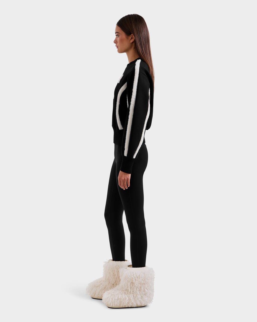 A woman in profile stands against a plain background, wearing après-ski chic in Rudsak’s CHANA - 8525656 Black boots—oversized with faux shearling trim—paired with a black outfit featuring white accents.