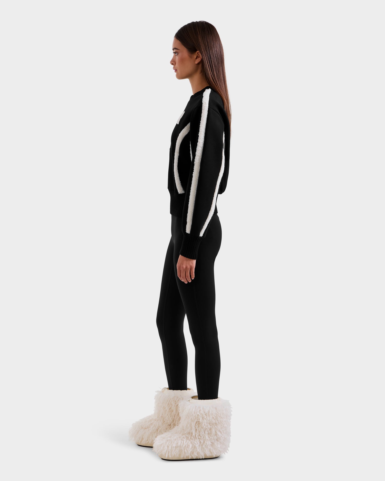 A woman in profile stands against a plain background, wearing après-ski chic in Rudsak’s CHANA - 8525656 Black boots—oversized with faux shearling trim—paired with a black outfit featuring white accents.