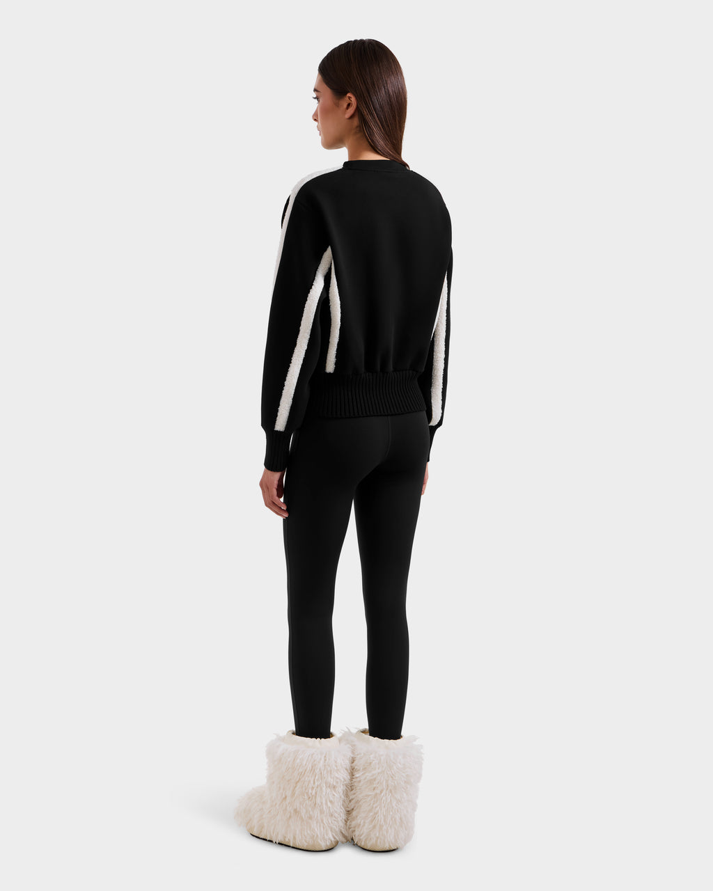 A woman faces away, wearing the Rudsak CHANA - 8525656 Black women's crew neck sweatshirt with white sleeve stripes, black leggings, and large white faux shearling trim boots. Her après-ski style stands out against a plain light gray backdrop.