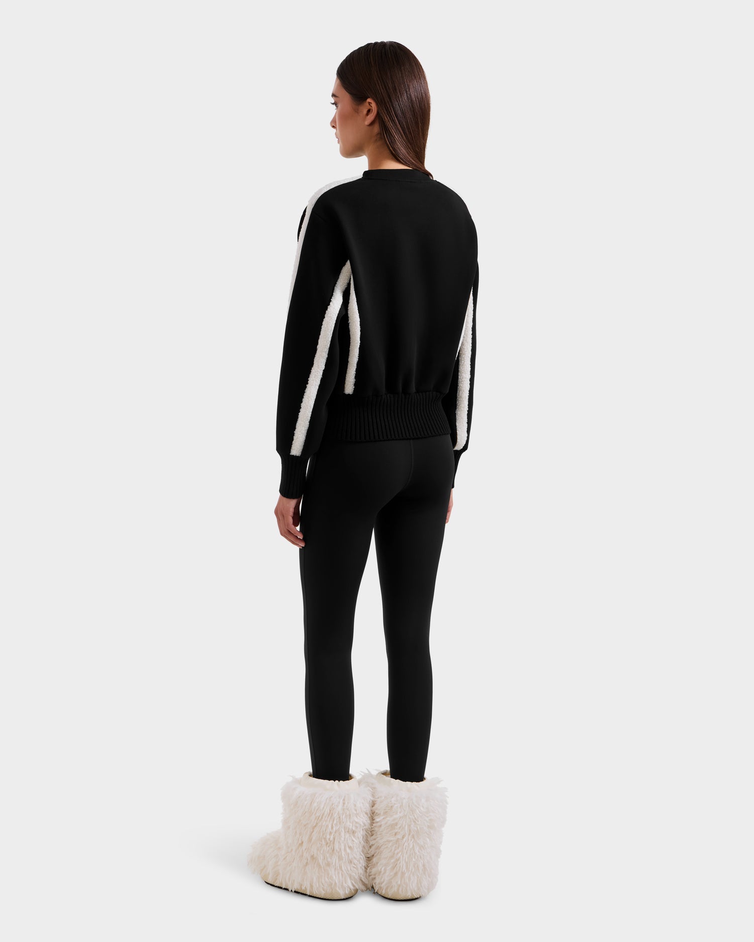 A woman faces away, wearing the Rudsak CHANA - 8525656 Black women's crew neck sweatshirt with white sleeve stripes, black leggings, and large white faux shearling trim boots. Her après-ski style stands out against a plain light gray backdrop.