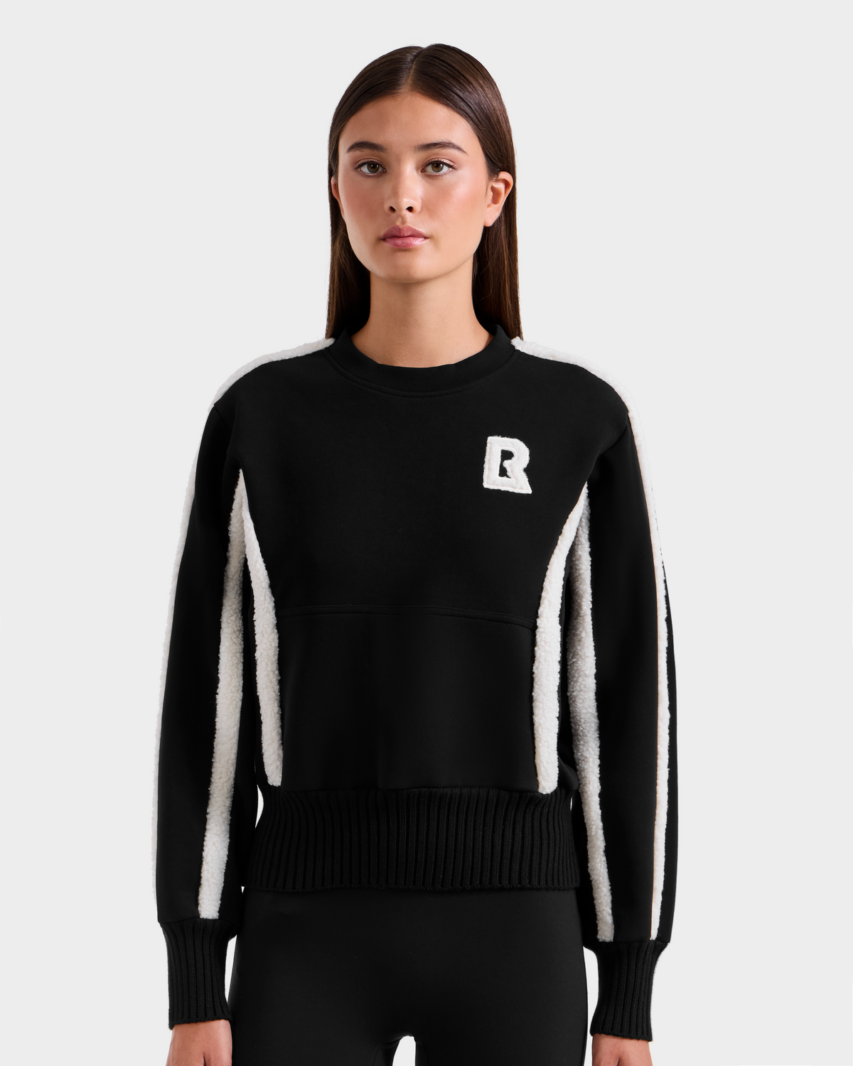 A young woman with straight brown hair wears the Rudsak CHANA - 8525656 Black women’s crew neck sweatshirt, featuring white accents and a bold white "R" on the chest, standing against a plain light gray background.
