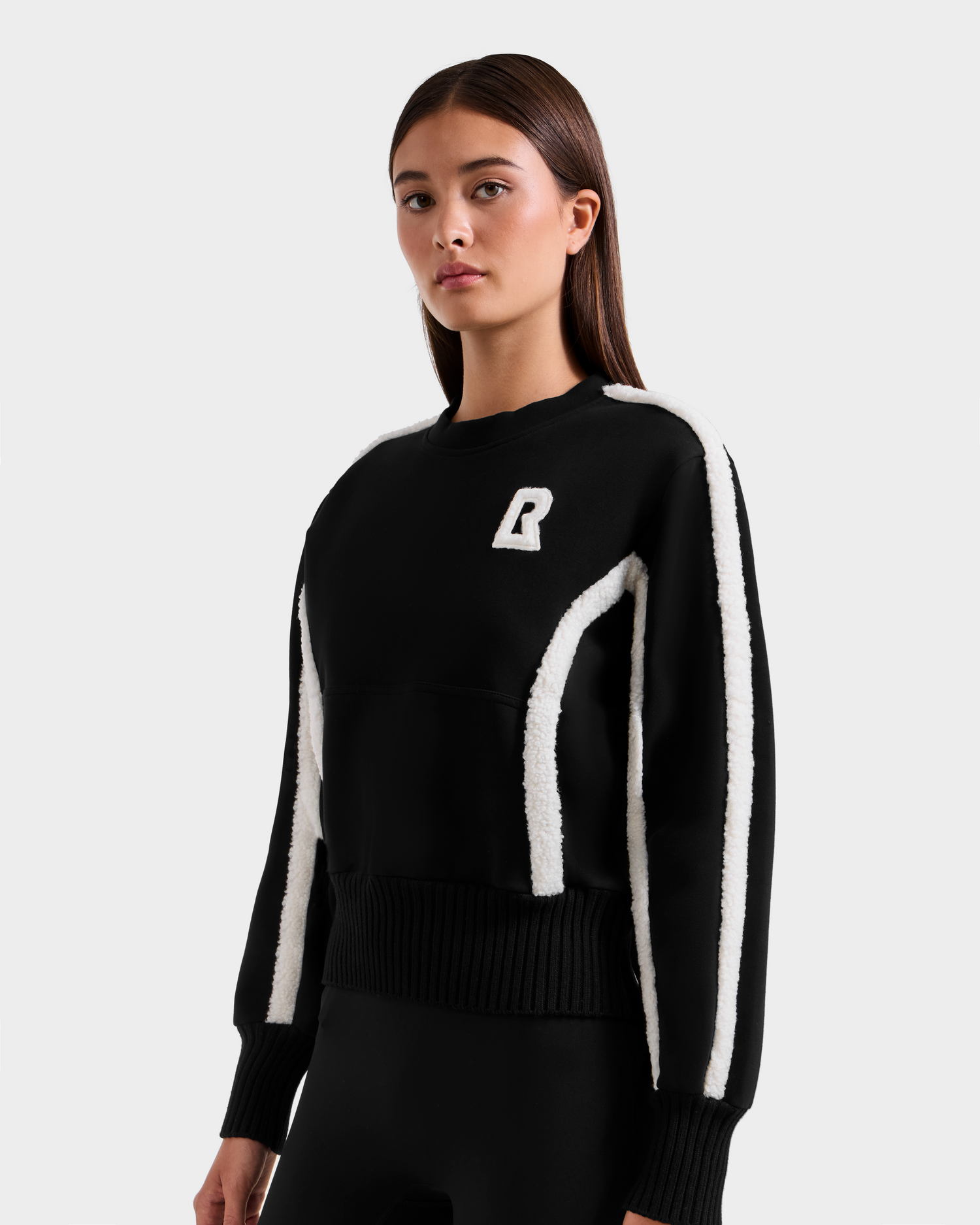 A woman with straight brown hair wears the Rudsak CHANA - 8525656 Black women's crew neck sweatshirt with white stripes and a white R logo, standing against a plain light background.