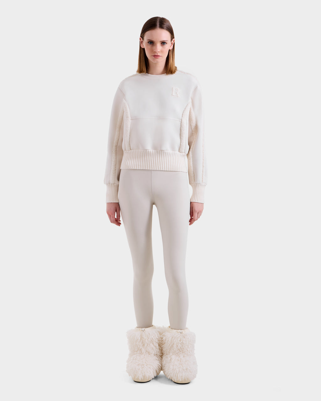 A woman faces forward in après-ski chic, wearing the Rudsak CHANA - 8525656 Vanilla crew neck sweatshirt with cream leggings and beige faux shearling-trim boots, set against a plain white background.