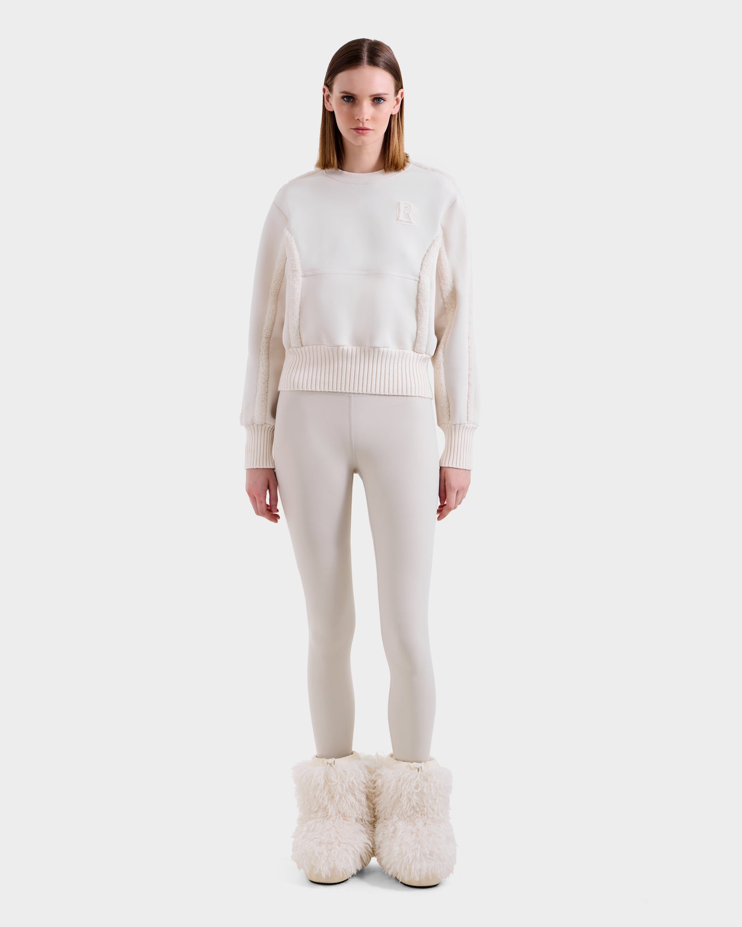 A woman faces forward in après-ski chic, wearing the Rudsak CHANA - 8525656 Vanilla crew neck sweatshirt with cream leggings and beige faux shearling-trim boots, set against a plain white background.