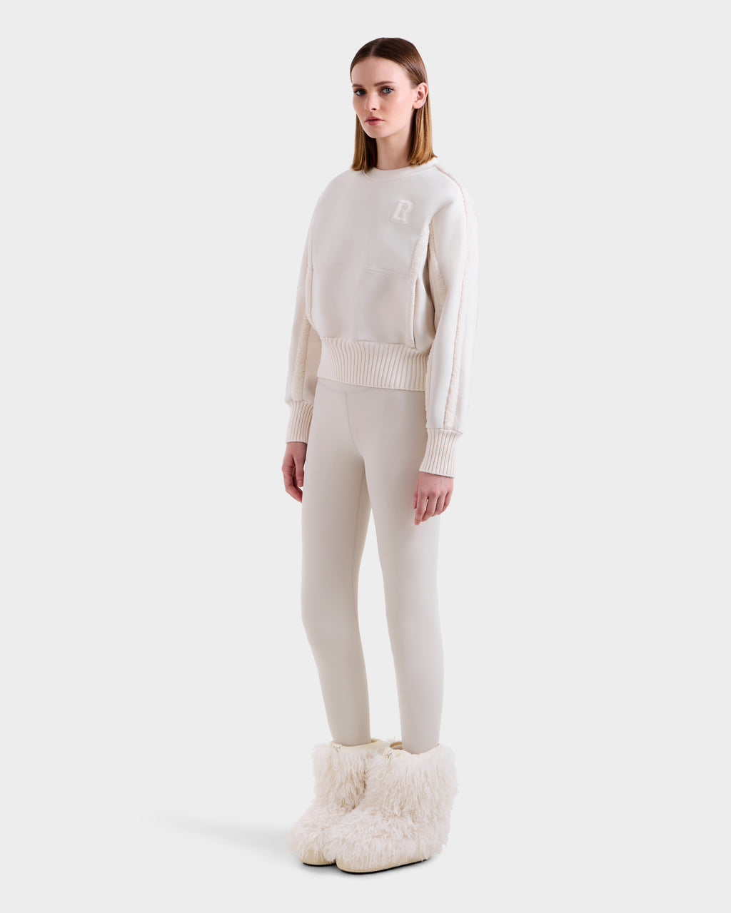 A woman poses against a plain background in the Rudsak CHANA - 8525656 Vanilla crew neck sweatshirt and matching leggings, paired with large faux shearling trim boots for a chic après-ski look. Her straight hair and neutral expression complete the style.