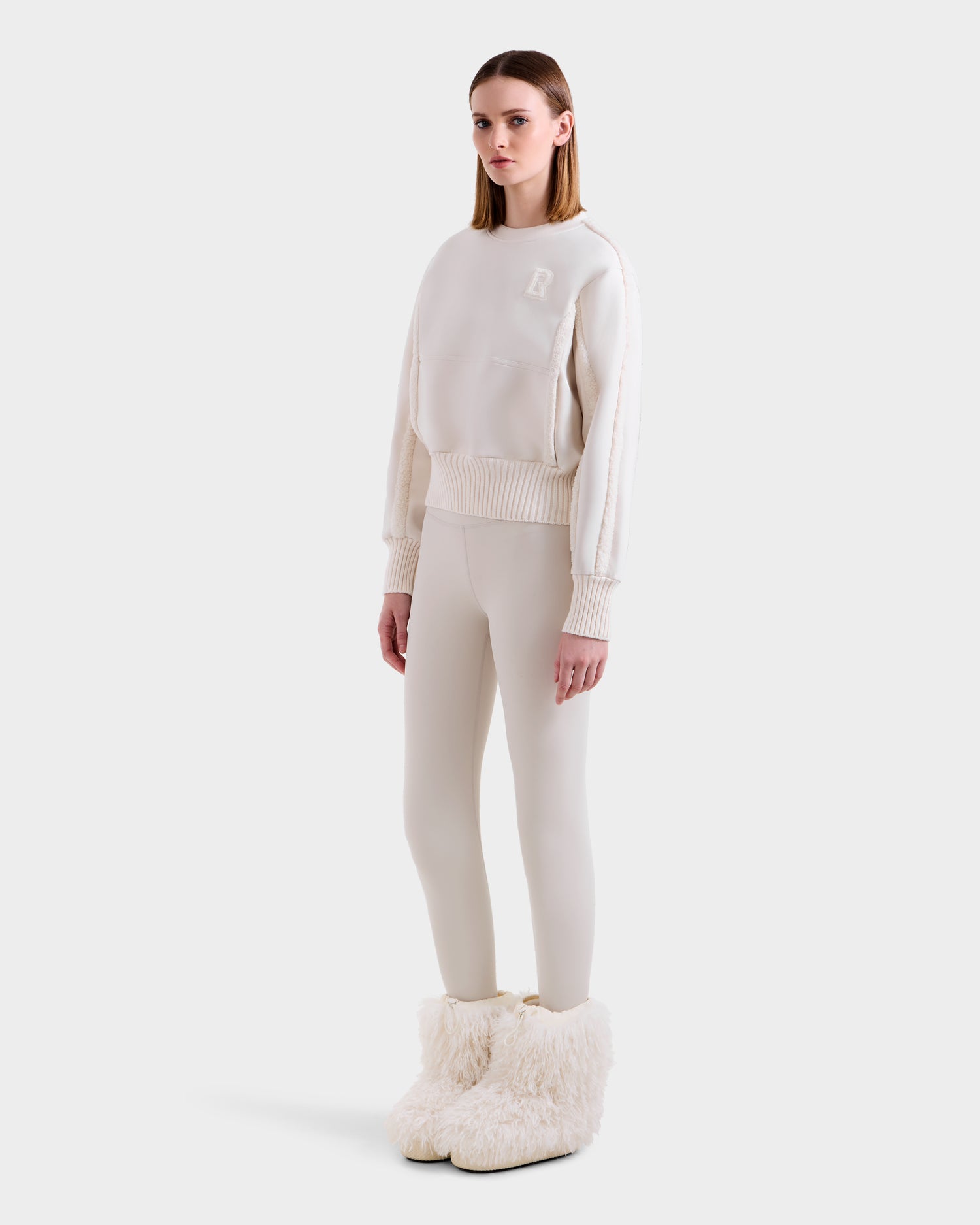 A woman poses against a plain background in the Rudsak CHANA - 8525656 Vanilla crew neck sweatshirt and matching leggings, paired with large faux shearling trim boots for a chic après-ski look. Her straight hair and neutral expression complete the style.
