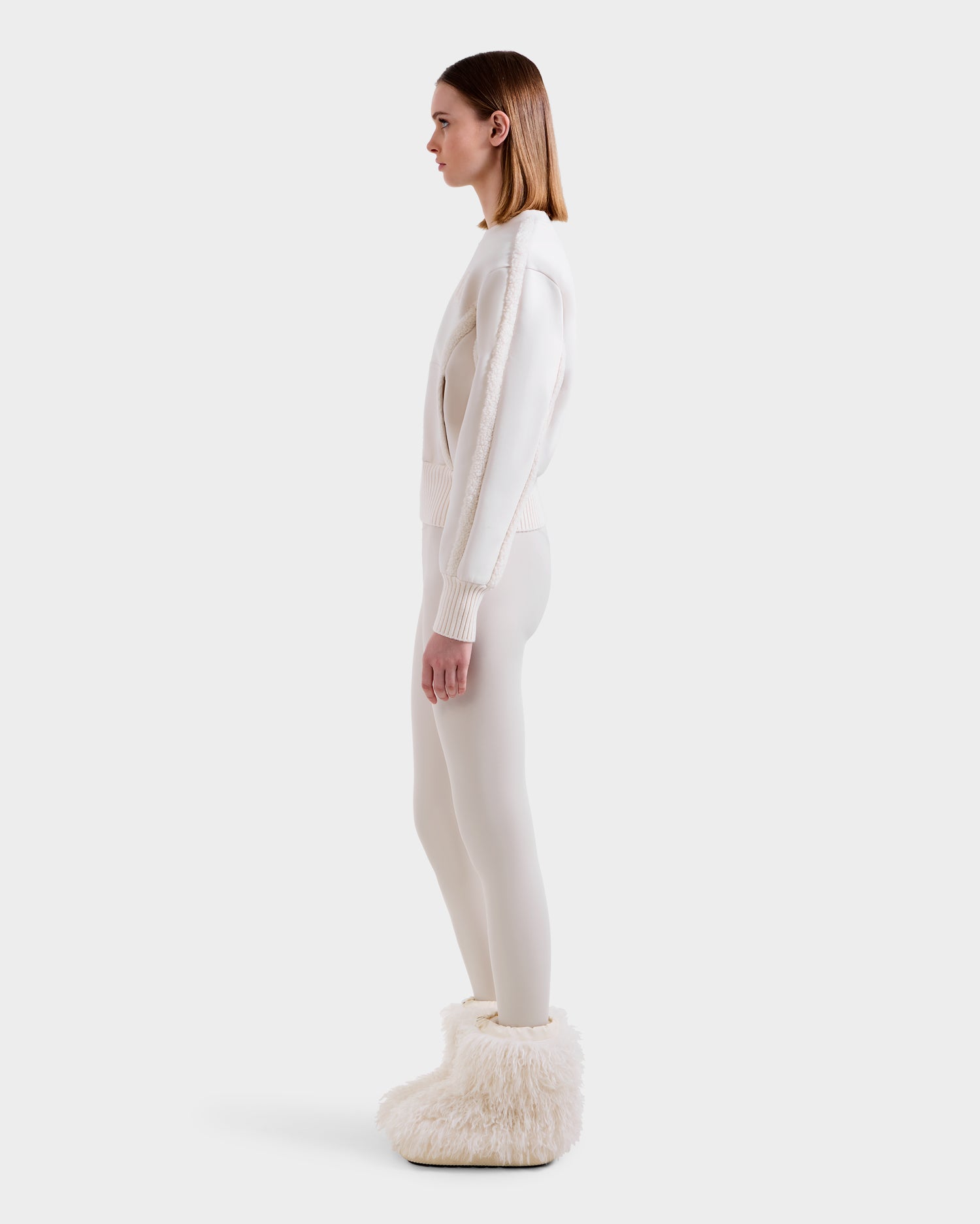 A woman in profile against a white background wears the Rudsak CHANA - 8525656 Vanilla crew neck sweatshirt with matching leggings and fluffy, faux shearling-trimmed white boots for an après-ski chic look.