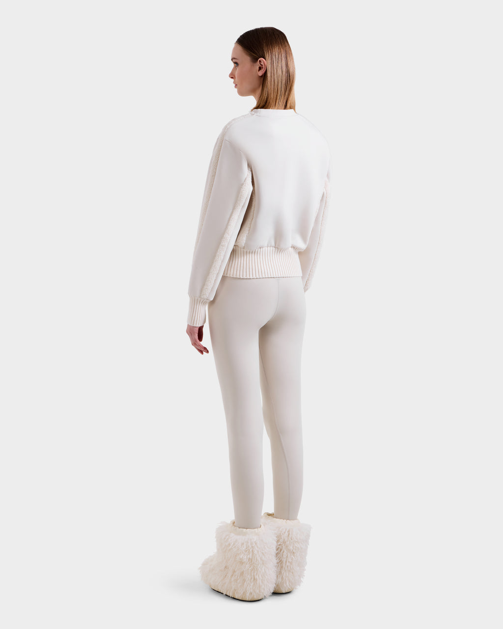 A woman stands facing away in après-ski chic style, wearing the Rudsak CHANA - 8525656 Vanilla women’s crew neck sweatshirt with white leggings and fluffy white boots against a plain white background.