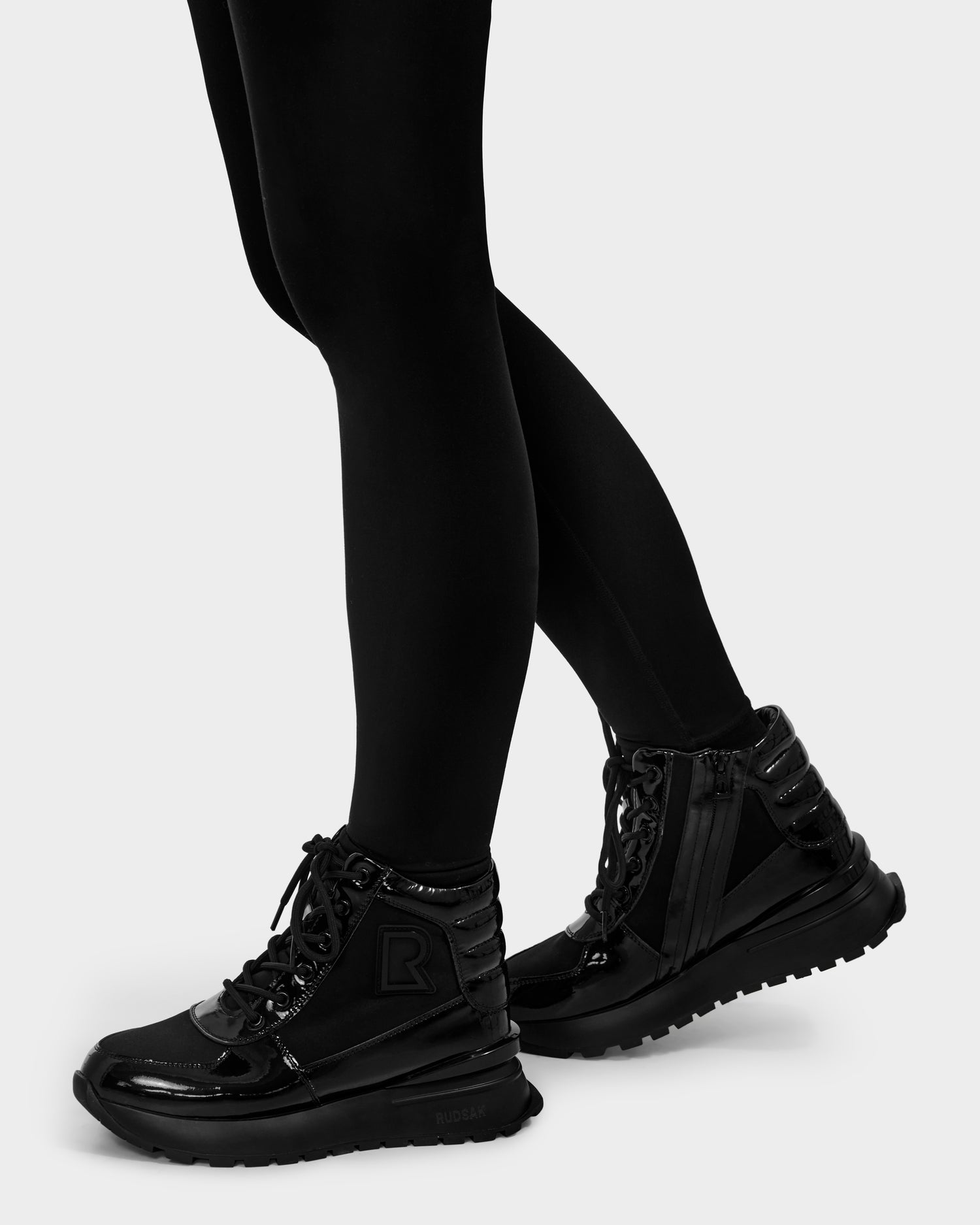 A person wearing Rudsak CHELSA - L224505 Black winter boots with thick soles and black leggings stands on a light gray background.