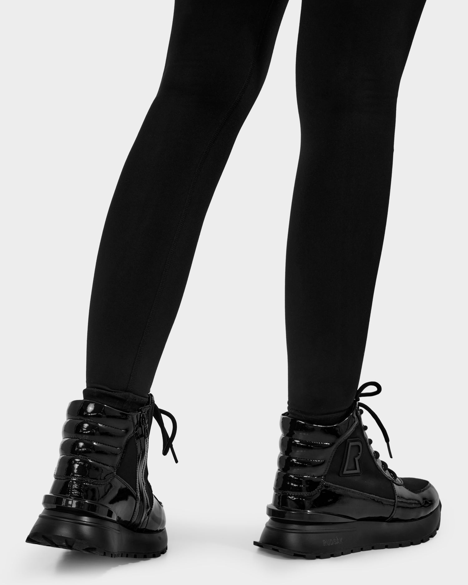 A person seen from behind at calf level wears Rudsak CHELSA - L224505 Black women's sneaker boots with a waterproof zipper and black leggings, set against a plain background.