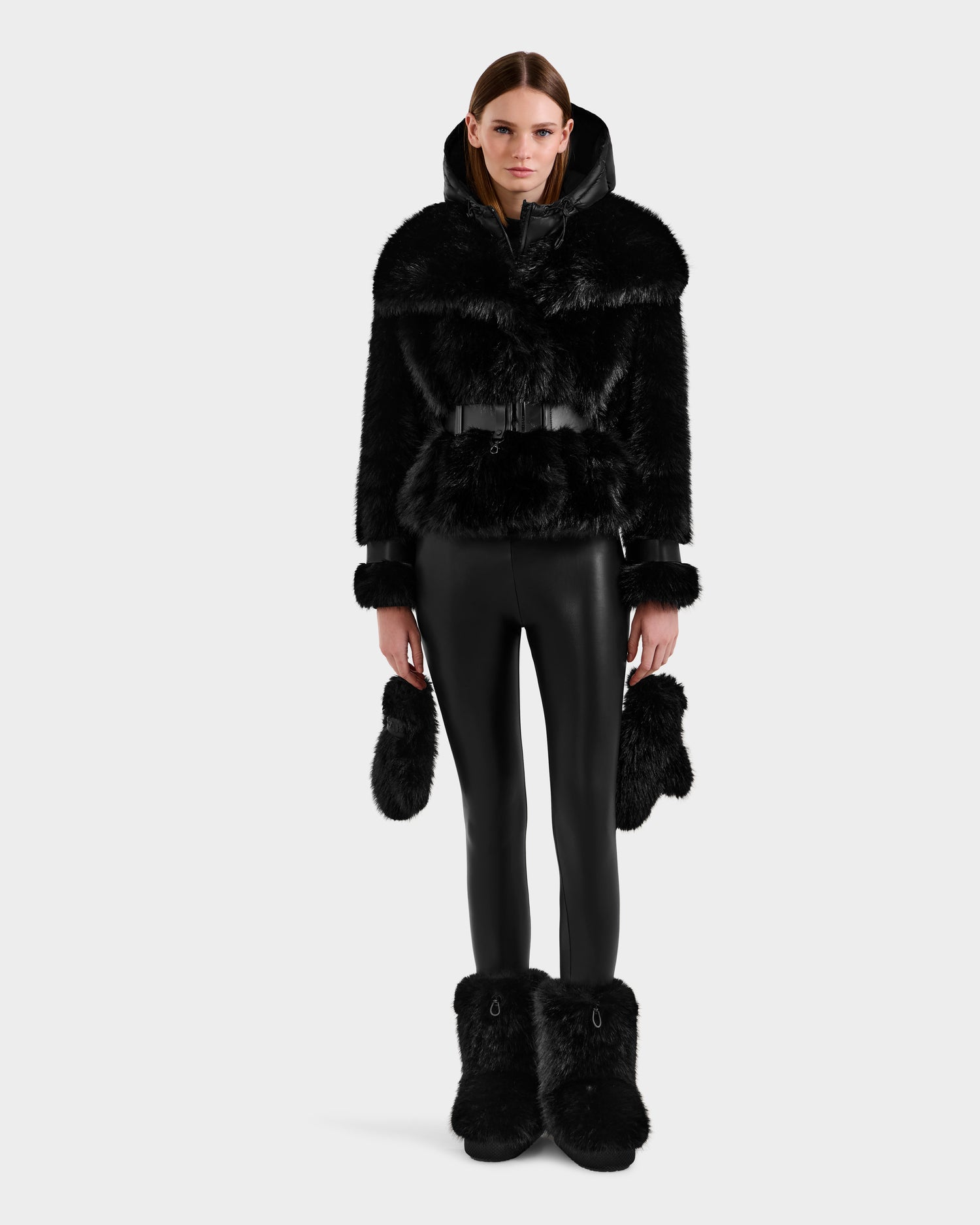 A woman faces forward in the Rudsak CHLOE - 8125619 Black faux fur jacket with belt, black vegan leather pants, matching faux fur cuffs and boots, holding a fur accessory. A plain white background highlights her chic winter look.