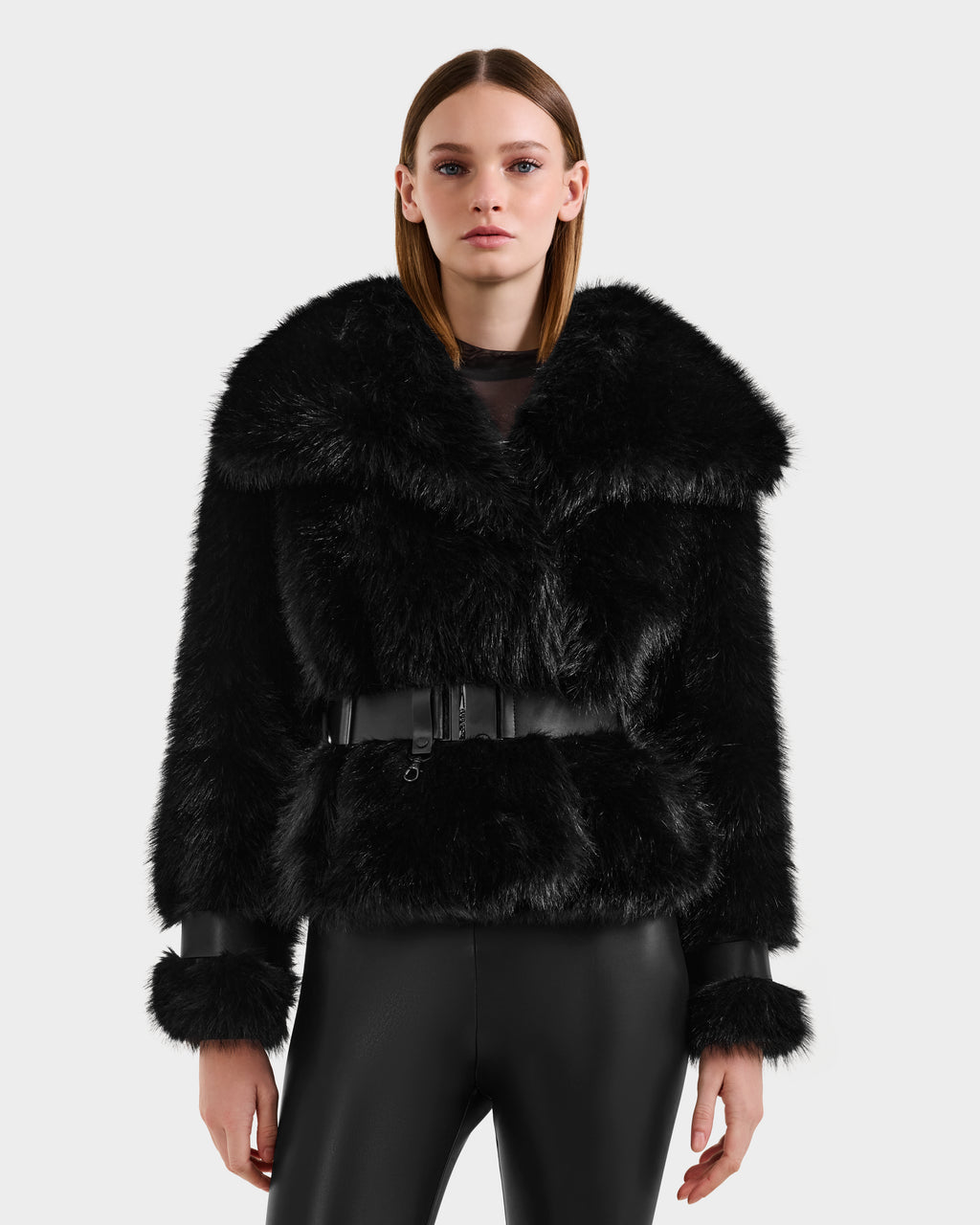 A woman with straight brown hair models the Rudsak CHLOE - 8125619 Black faux fur jacket, featuring a wide collar, black belt, and leather-trimmed cuffs, paired with black leather pants against a simple light background.
