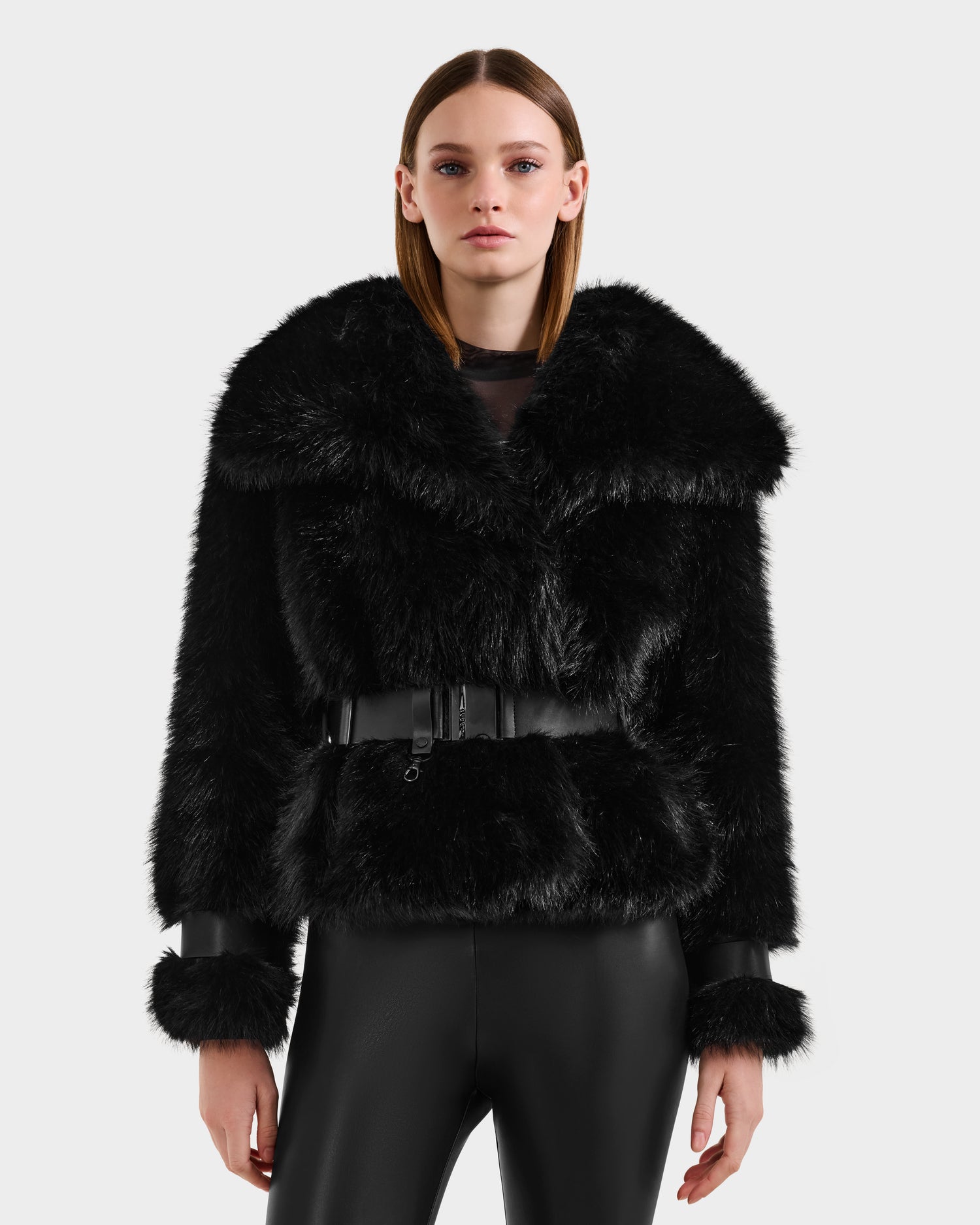 A woman with straight brown hair models the Rudsak CHLOE - 8125619 Black faux fur jacket, featuring a wide collar, black belt, and leather-trimmed cuffs, paired with black leather pants against a simple light background.