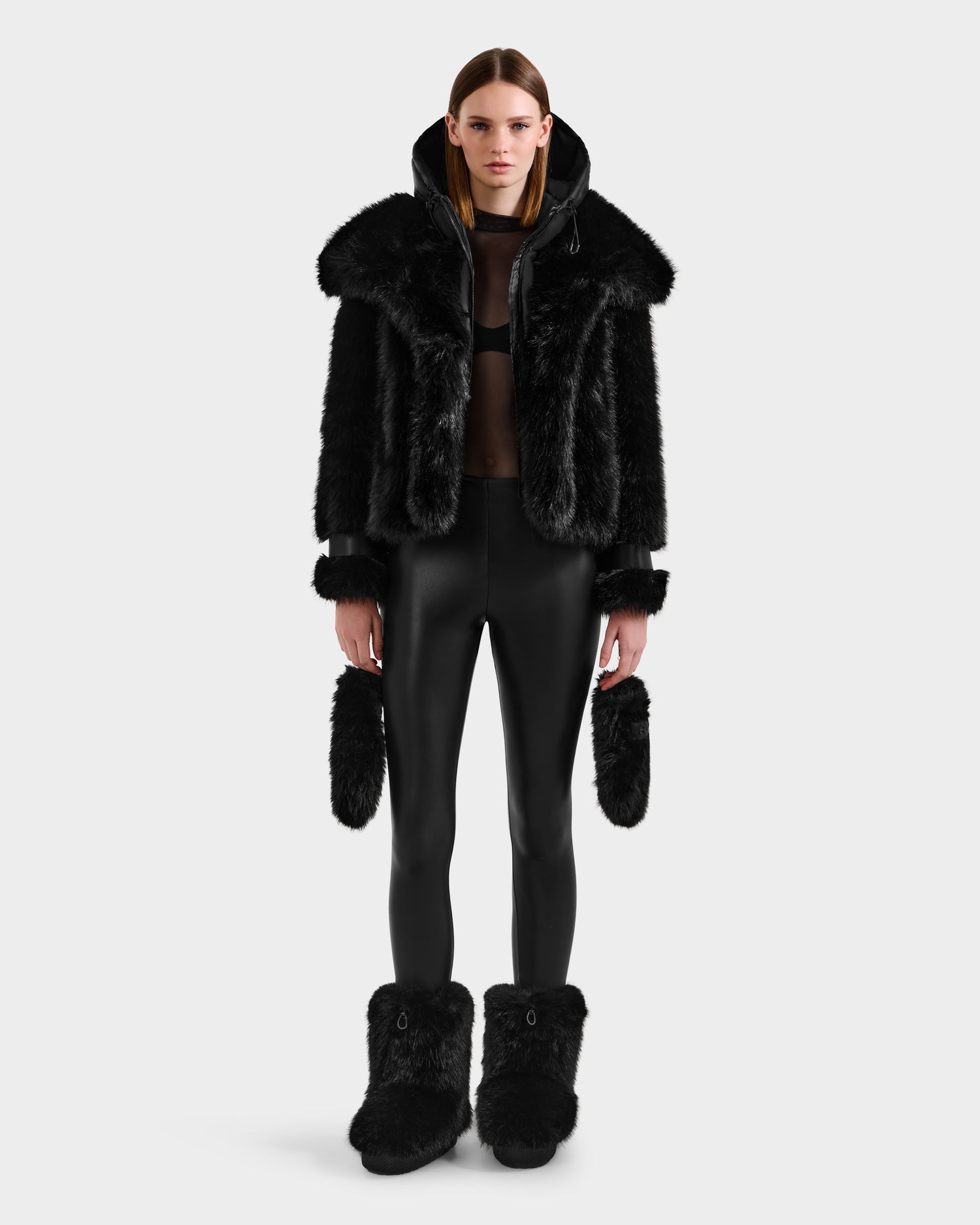 A person stands facing forward against a white background, wearing the Rudsak CHLOE - 8125619 Black women’s faux fur jacket with a hood—an effortlessly chic choice for your winter wardrobe.