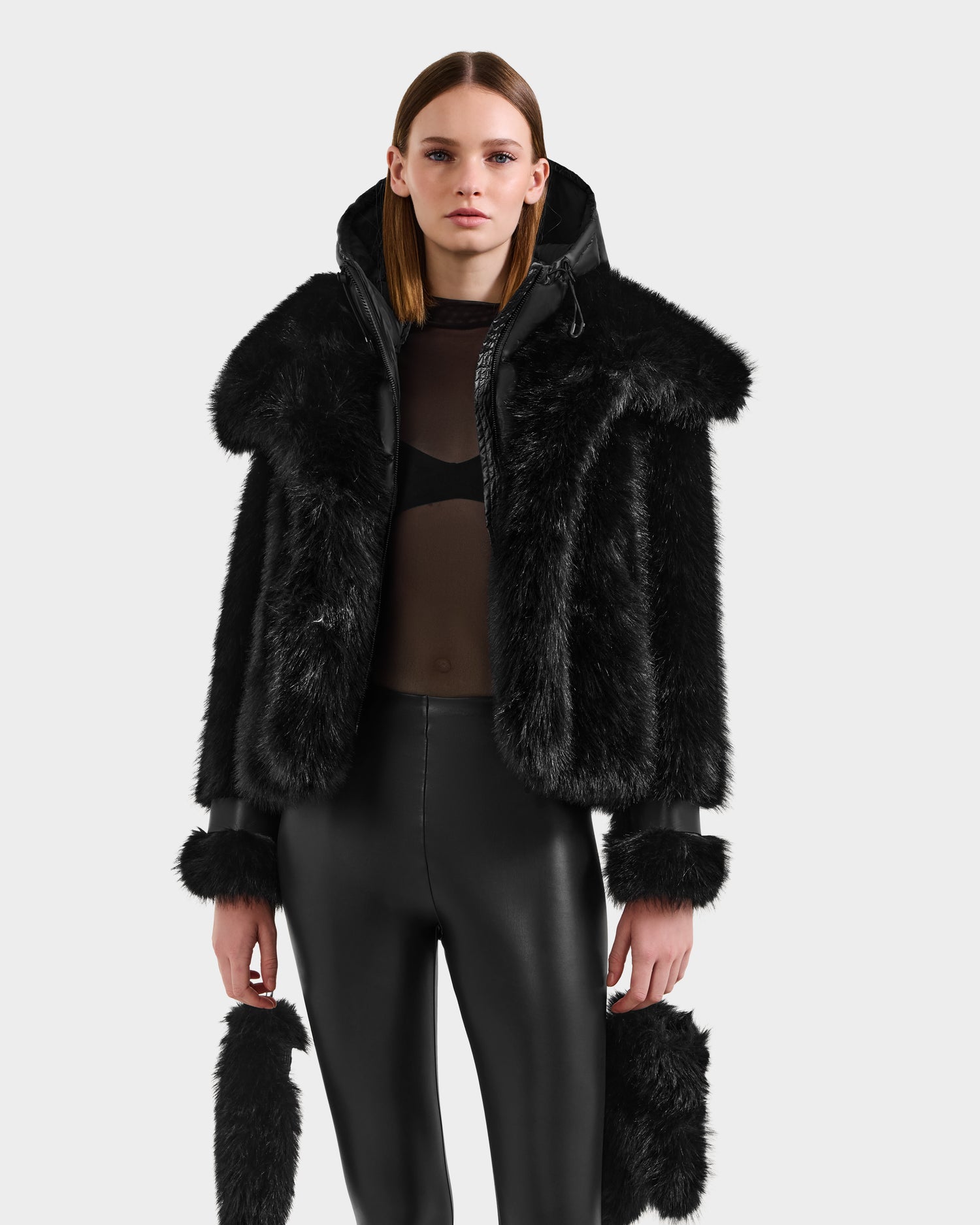 A woman wears the Rudsak CHLOE - 8125619 Black faux fur jacket with a large collar and cuffs over a sheer black top and high-waisted leggings, standing against a plain light background—perfect for your winter wardrobe.