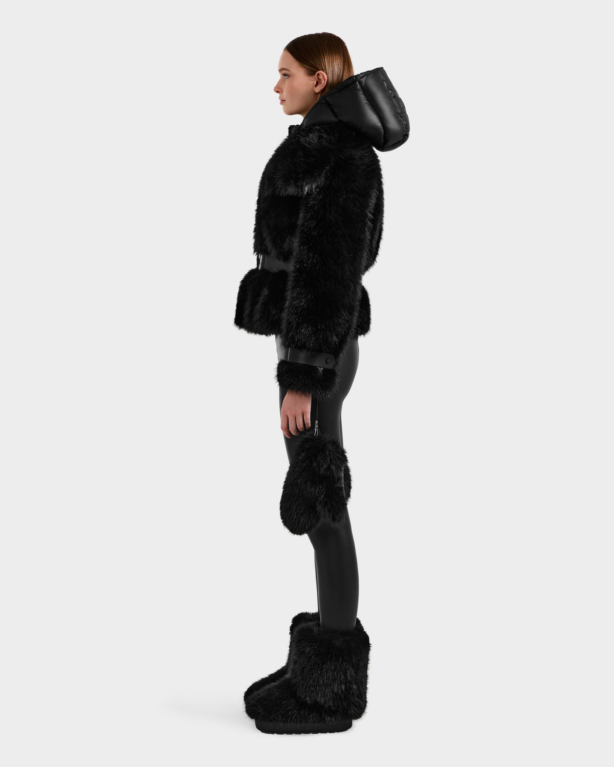 A woman stands in profile against a white background wearing the Rudsak CHLOE - 8125619 Black faux fur jacket with a hood, black leggings, furry leg warmers, and black furry boots—a chic winter fashion look.