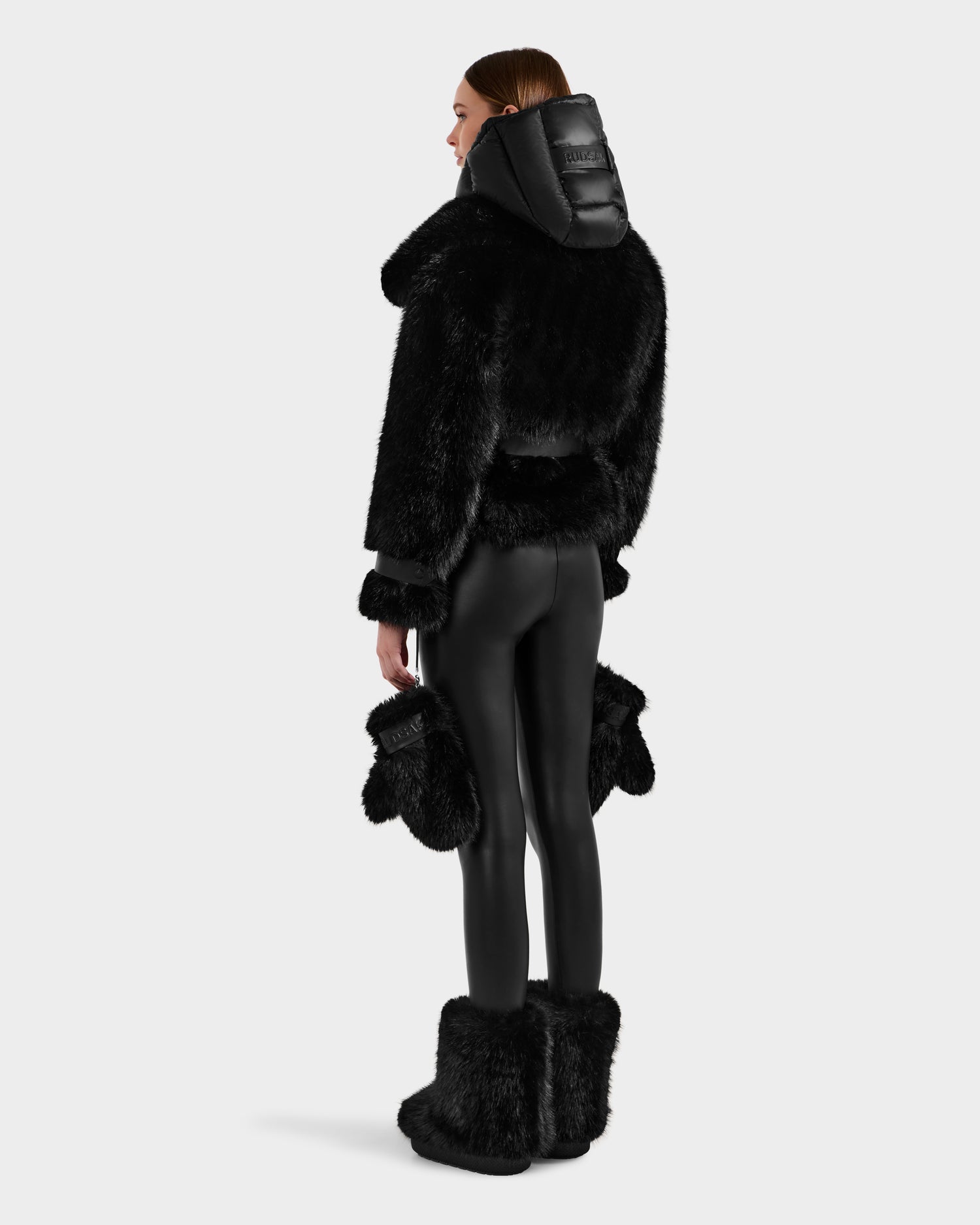 A woman faces away, modeling the Rudsak CHLOE - 8125619 Black faux fur jacket with a large hood, black fitted pants, and matching black faux fur boots and mittens against a plain light background.