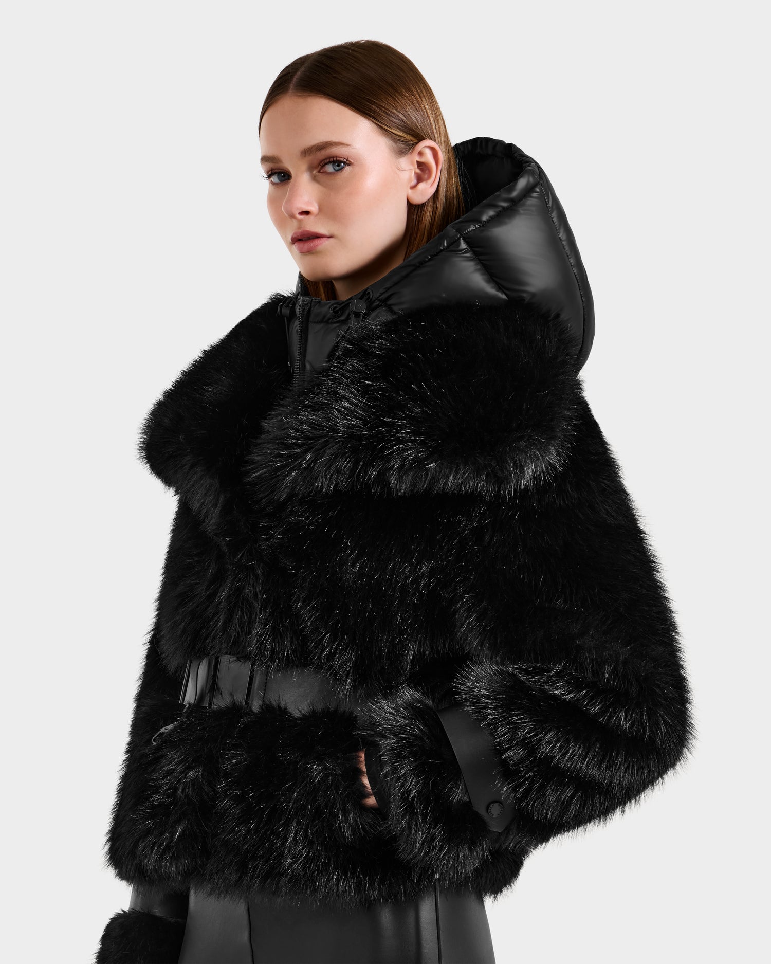 A woman looks to the side in the Rudsak CHLOE - 8125619 Black faux fur jacket, featuring a shiny hood, plush faux fur trim, and vegan leather accents on the body and cuffs, set against a plain light gray background.