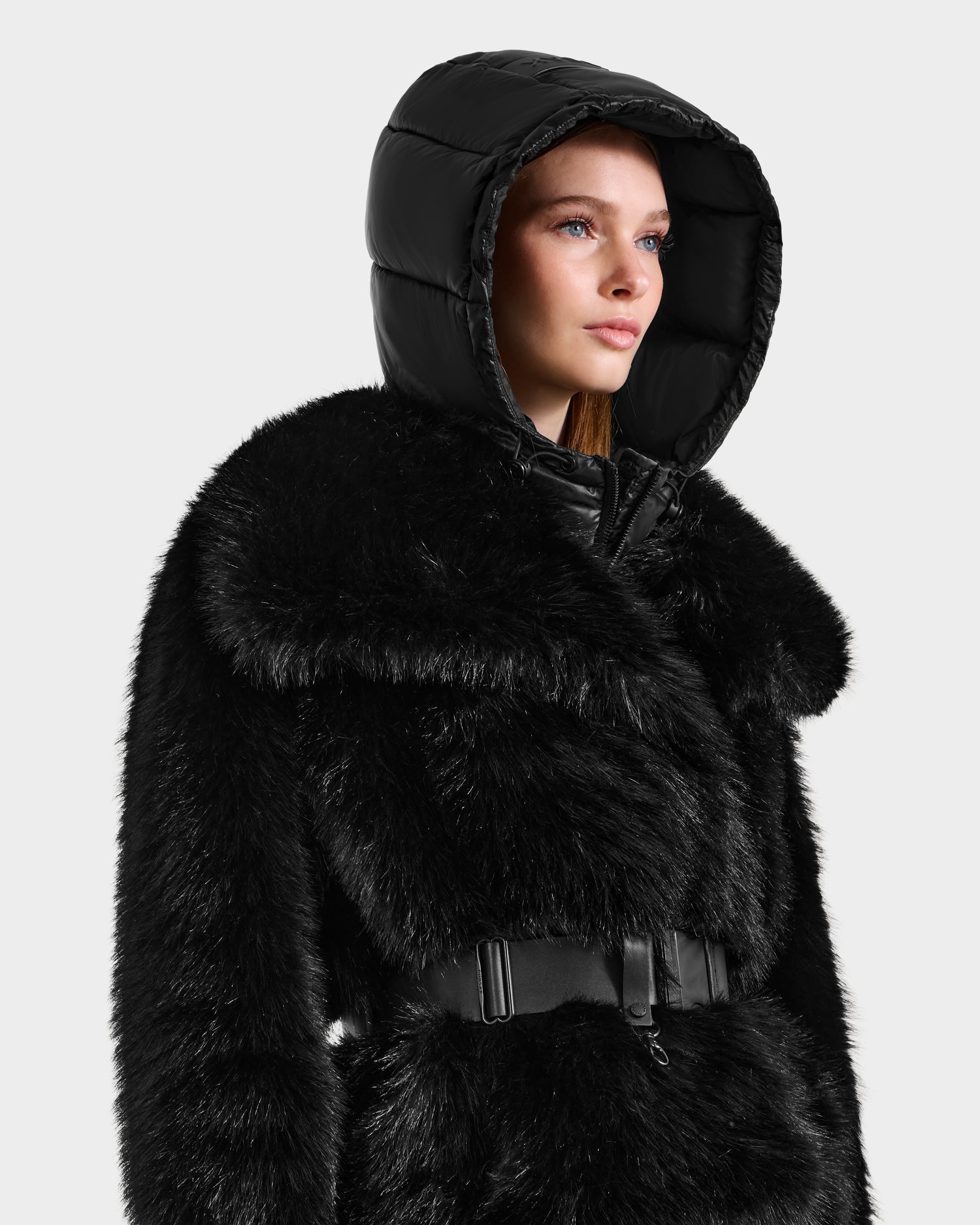 A woman models the Rudsak CHLOE - 8125619 Black women's faux fur jacket with a wide belt and shiny black hood, looking aside against a white background—an ideal winter fashion choice.