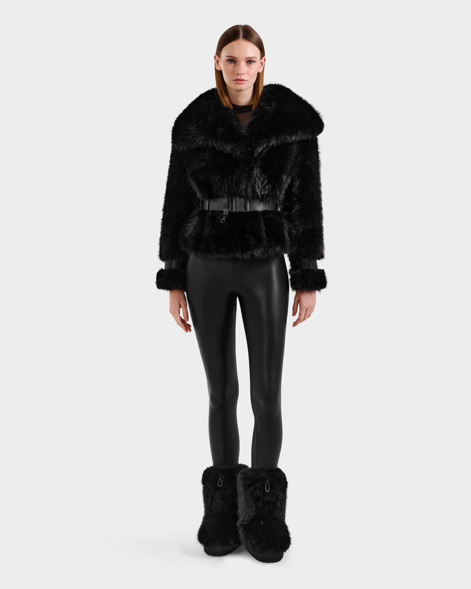 A woman faces forward wearing the CHLOE - 8125619 Black faux fur jacket by Rudsak, paired with black faux fur boots and shiny leggings—a chic winter look set against a plain light background.