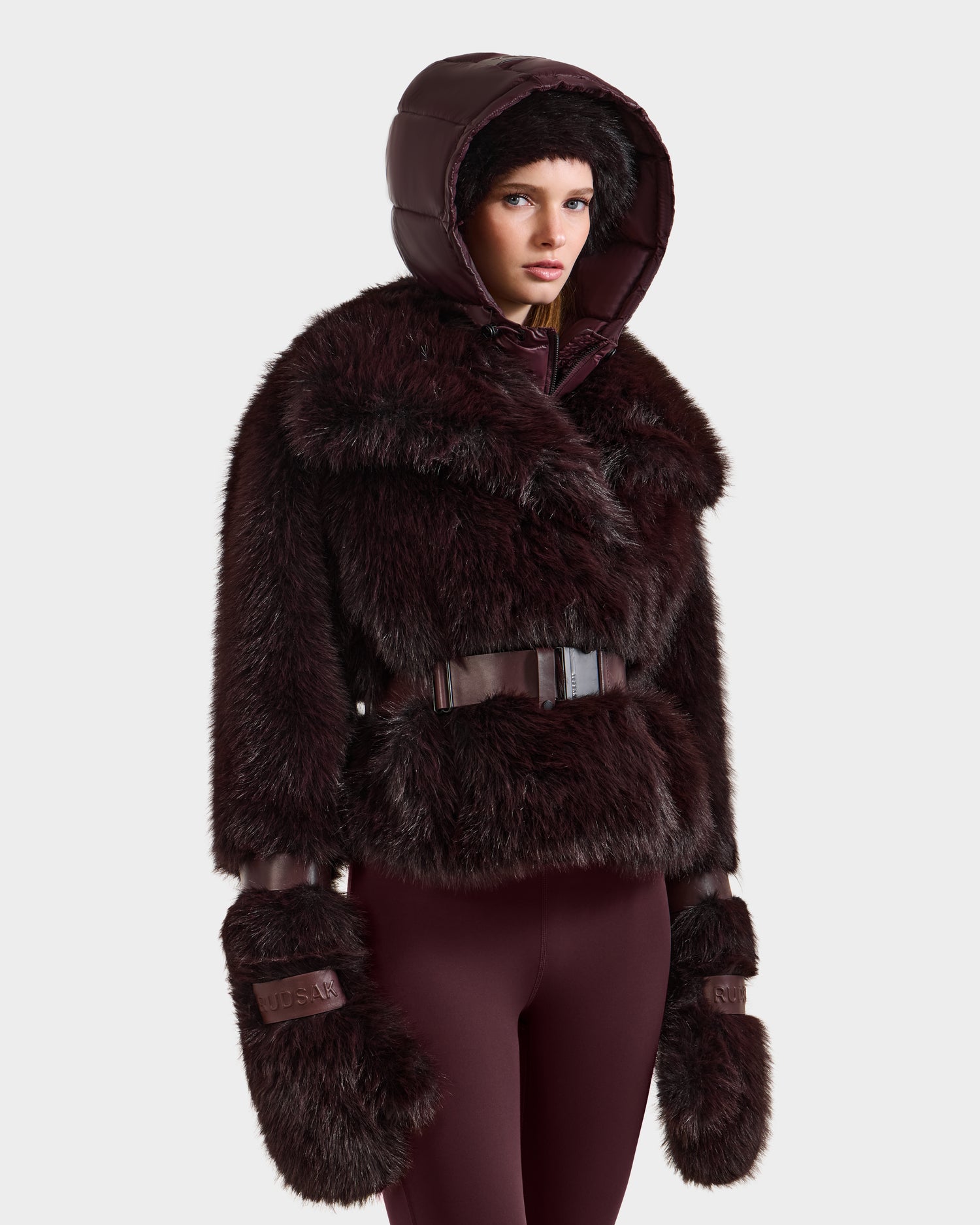 A woman wears Rudsak's CHLOE - 8125619 Bordeaux faux fur jacket with a hood, matching mittens, and burgundy leggings against a light gray background. The belted jacket features glossy accents for a chic winter look.